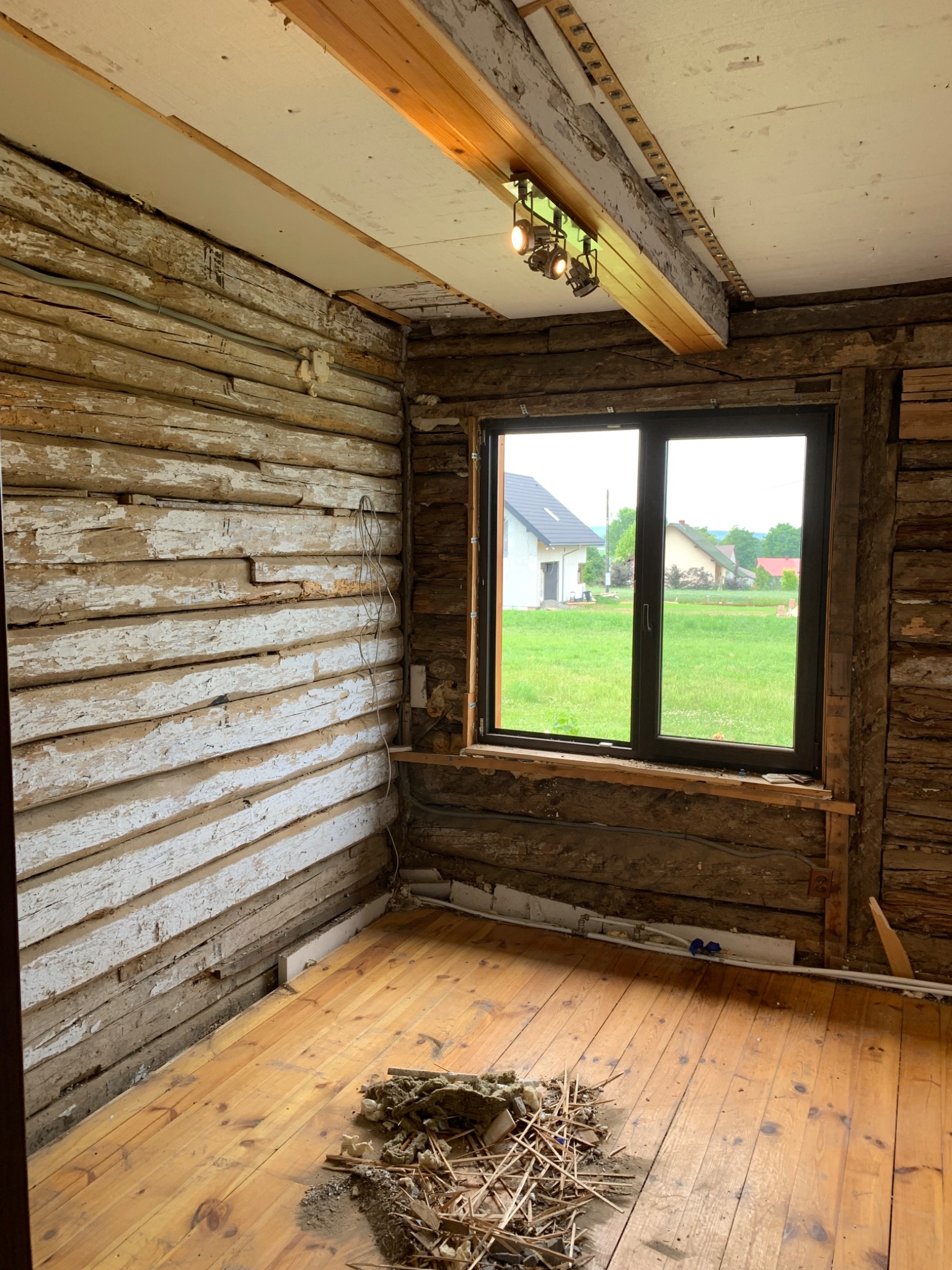 Wnętrze pokoju w trakcie remontu: ściany z drewnianych bali z resztkami białej farby, drewniana podłoga, okno z widokiem na domy, widoczne instalacje elektryczne i oświetlenie sufitowe.