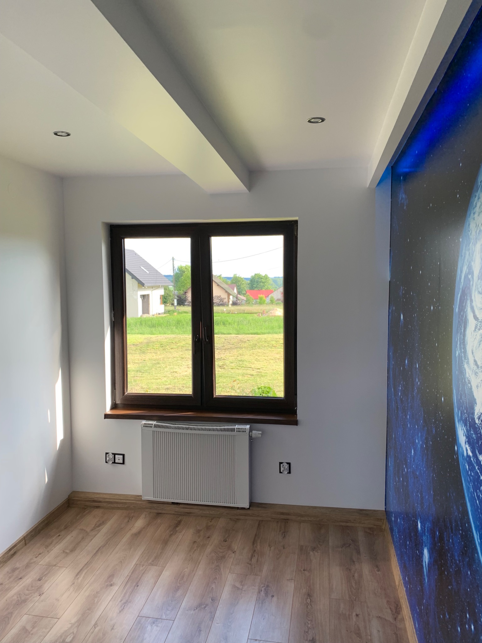 Jasne wnętrze pokoju po remoncie z drewnopodobną podłogą, grzejnikiem pod oknem i fototapetą z motywem kosmosu na ścianie.