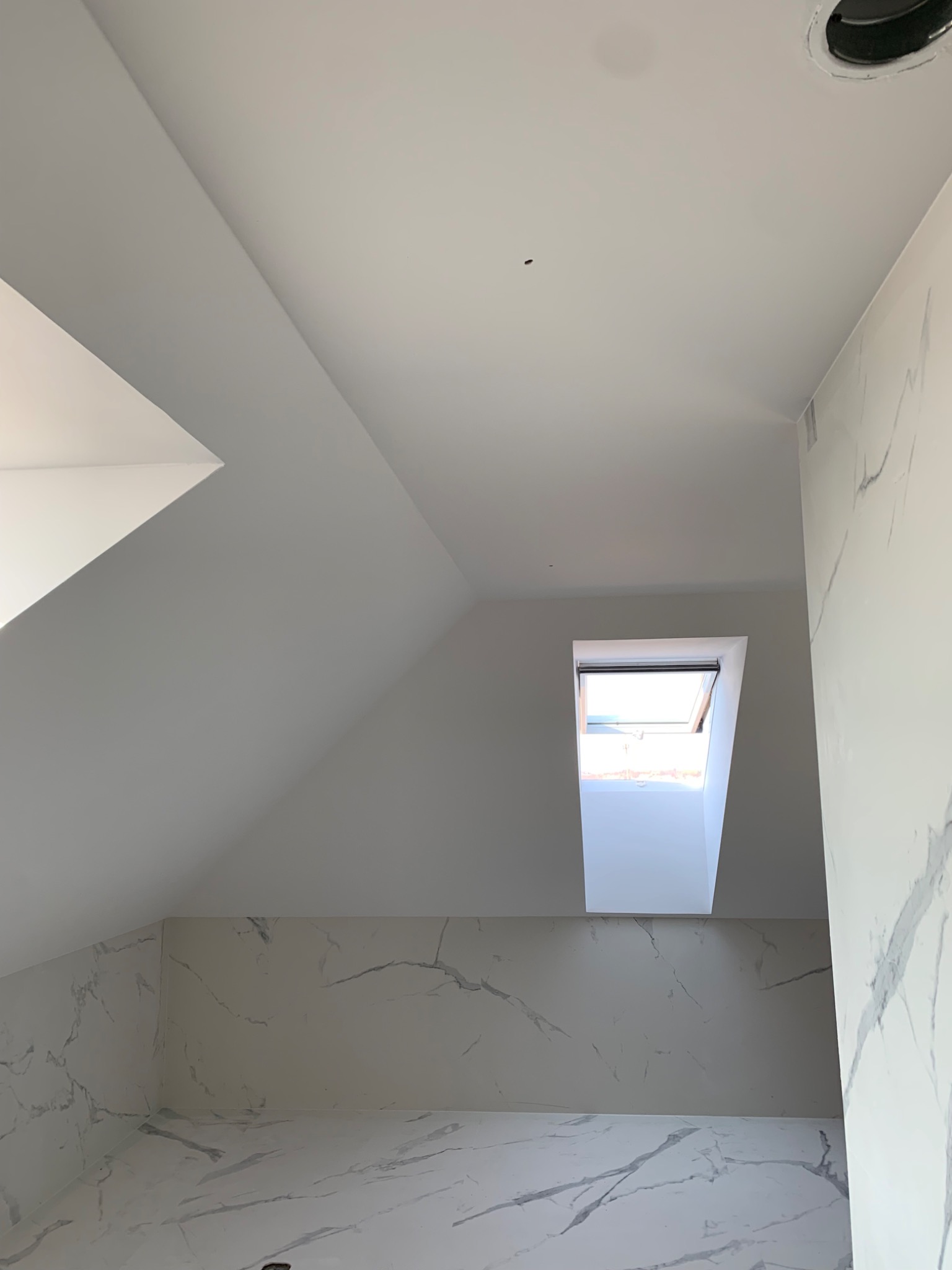Poddasze po remoncie z oknem dachowym, ściany wykończone płytami imitującymi marmur, biały sufit z widocznym otworem wentylacyjnym.