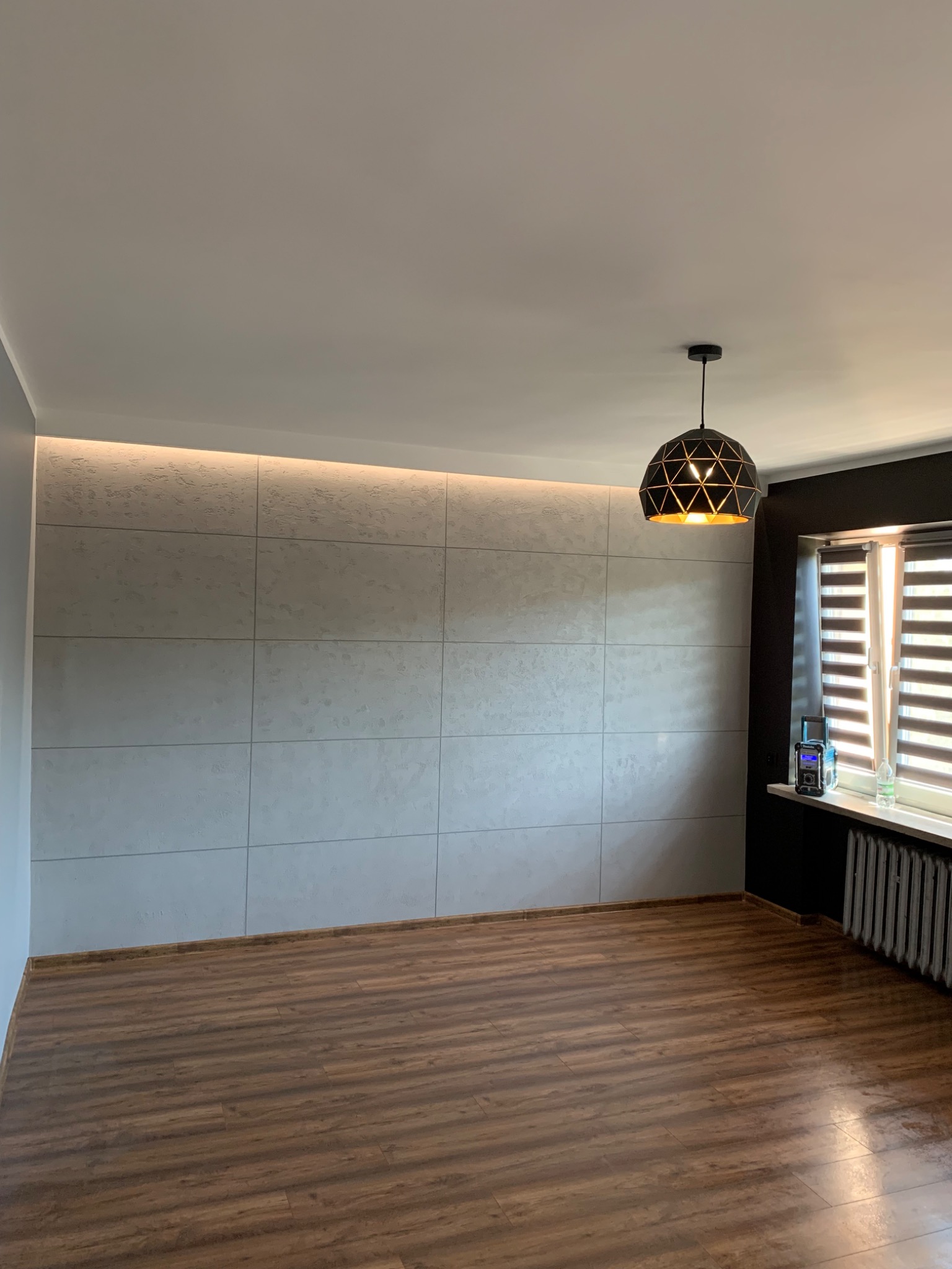 Pokój po remoncie: ściana wykończona płytami imitującymi beton z podświetleniem LED, drewnopodobna podłoga, czarna ściana z oknem i grzejnikiem, nowoczesna lampa wisząca.