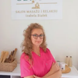 Portret kobiety w różowym uniformie na tle gabinetu z logo 'Odnowa Salon Masażu i Relaksu Izabela Białek', obok bambusowe kije i podgrzewacz do olejków.