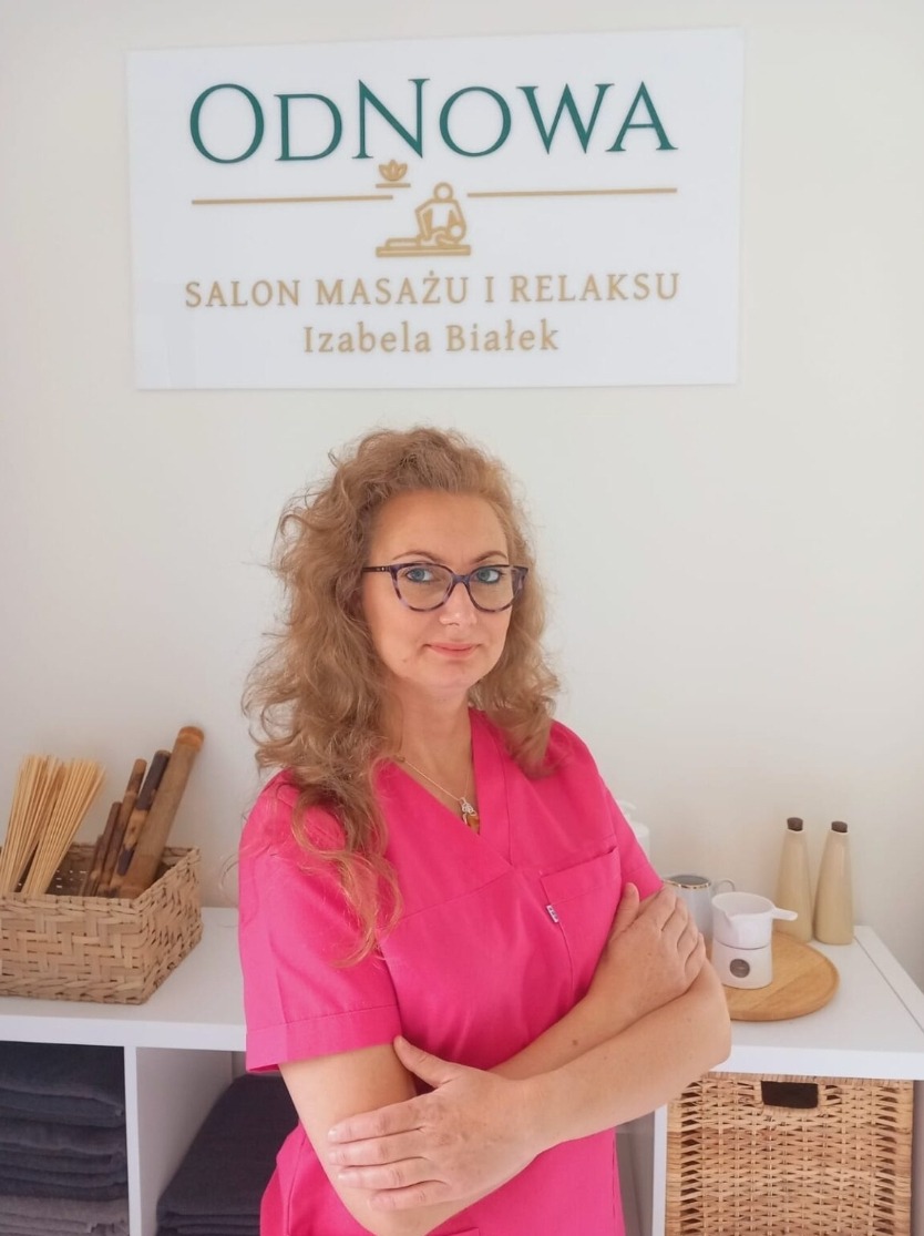 Portret kobiety w różowym uniformie na tle gabinetu z logo 'Odnowa Salon Masażu i Relaksu Izabela Białek', obok bambusowe kije i podgrzewacz do olejków.