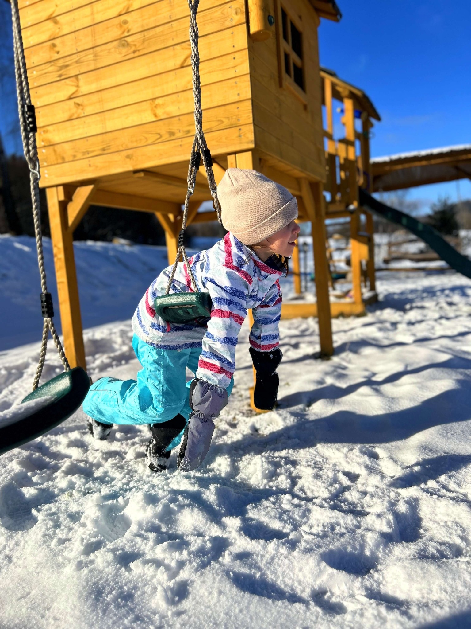 Dziecko w czapce i zimowych ubraniach bawi się na huśtawce linowej przed drewnianym placem zabaw w zimowej scenerii, z widocznym śniegiem i niebieskim niebem w tle.