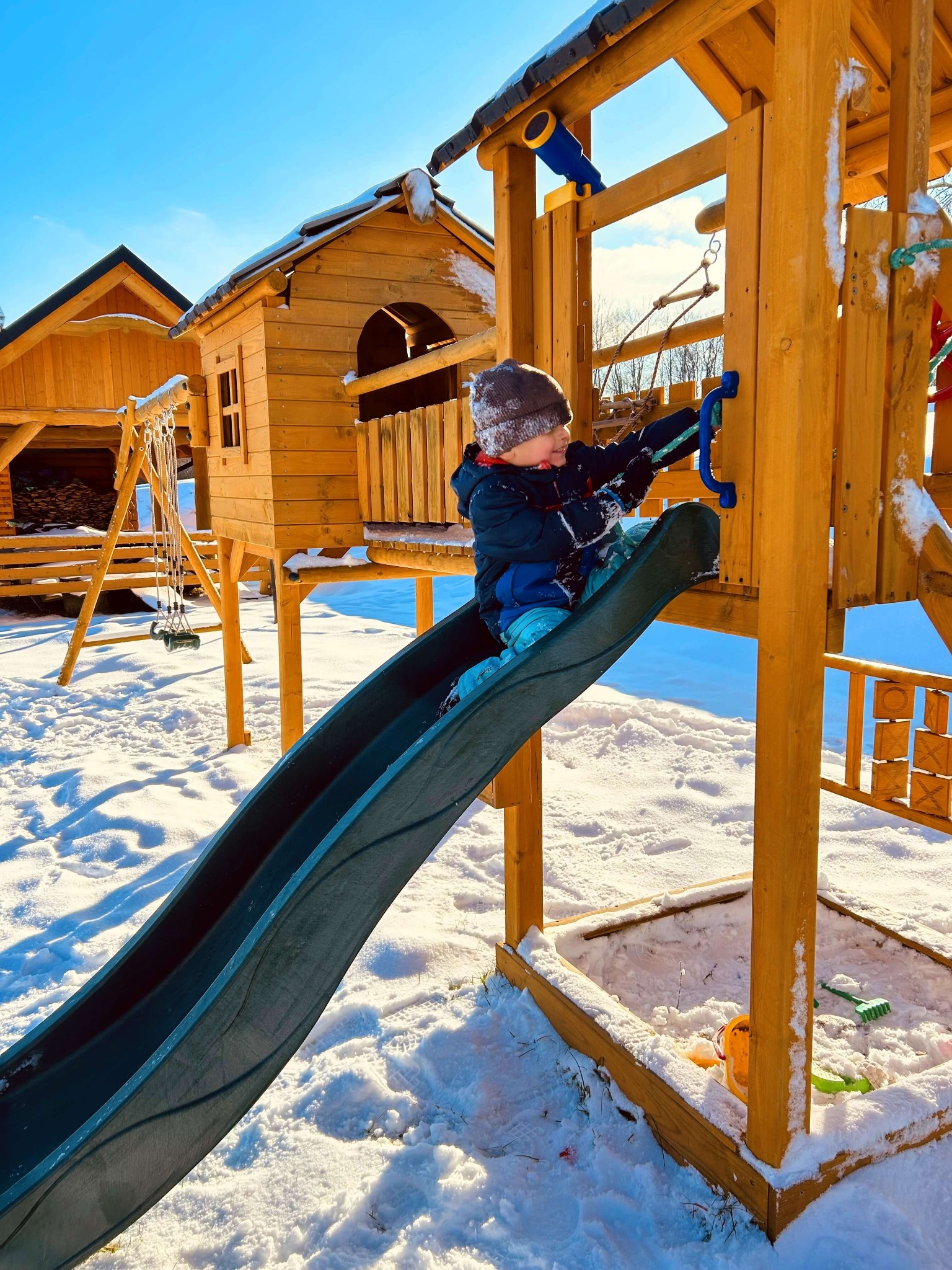 Dziecko wspina się po zjeżdżalni na drewnianym placu zabaw w zimowej scenerii, z domkiem na palach i huśtawką w tle, całość pokryta śniegiem w słoneczny dzień.