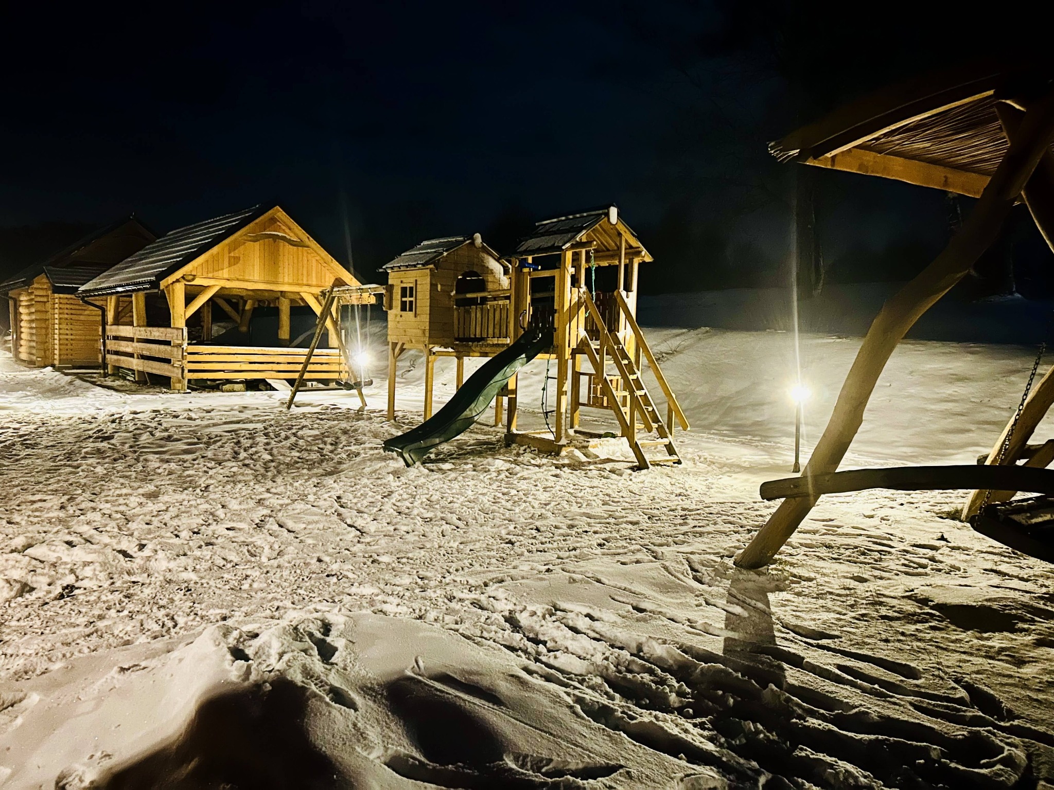 Oświetlony plac zabaw ze zjeżdżalnią i domkami dla dzieci, pokryty śniegiem, w nocy.