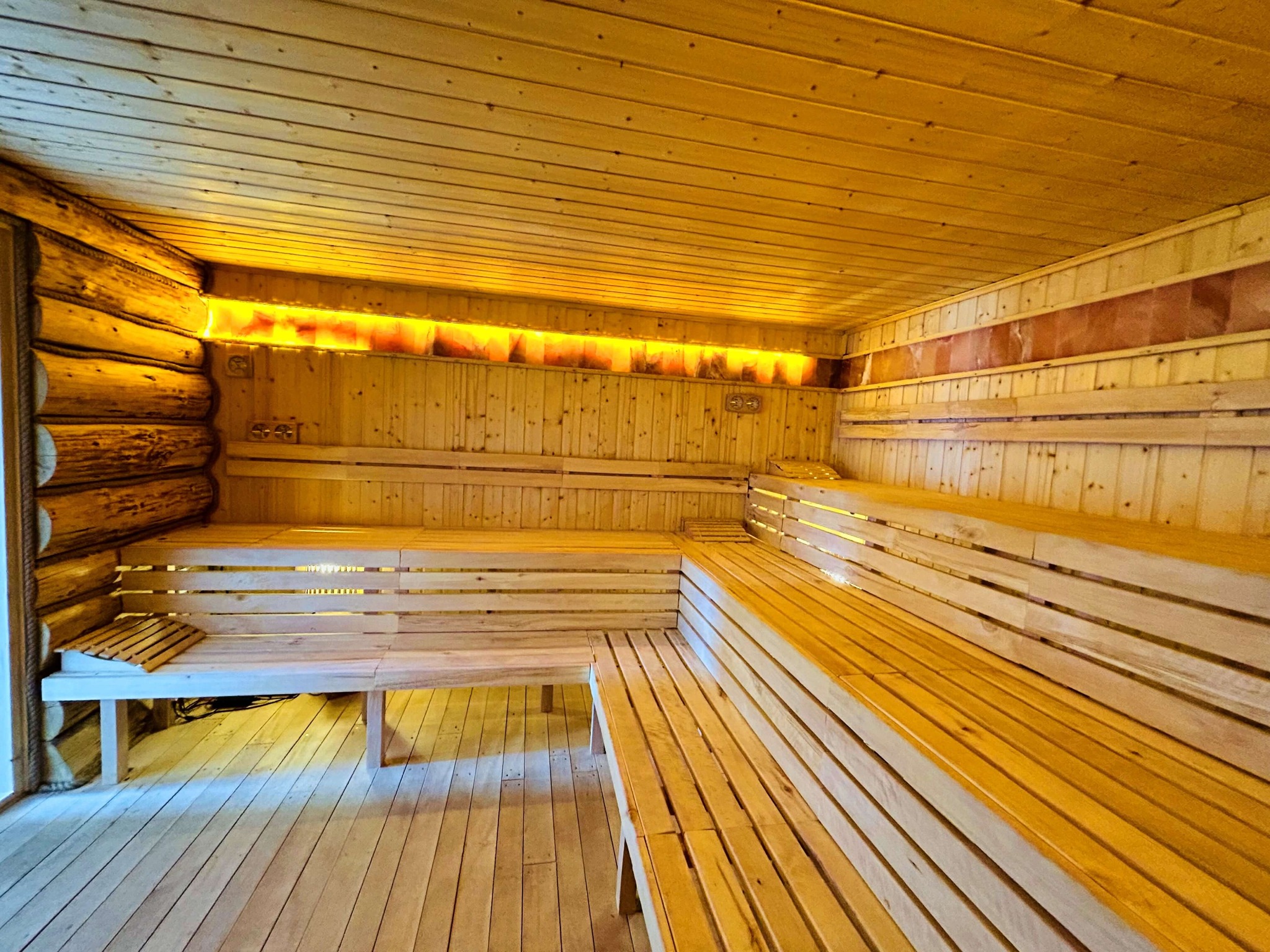 Wnętrze sauny wyłożone jasnym drewnem, z ławkami i podświetlanymi panelami solnymi w tle. Drewniana podłoga i ściany tworzą ciepły, relaksujący klimat.