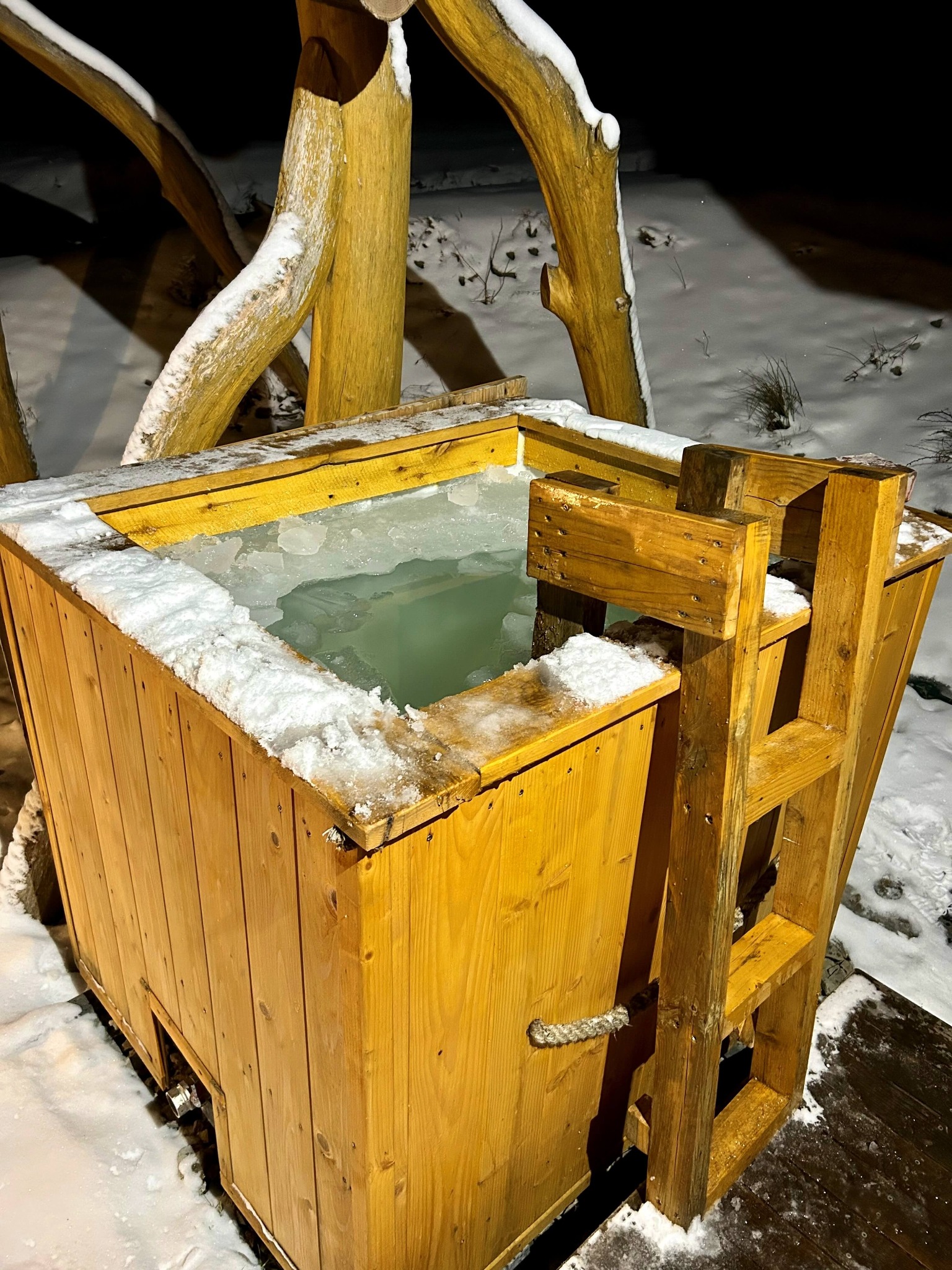 Drewniana balia z drabinką, wypełniona wodą z krą, otoczona śniegiem, oświetlona nocą.