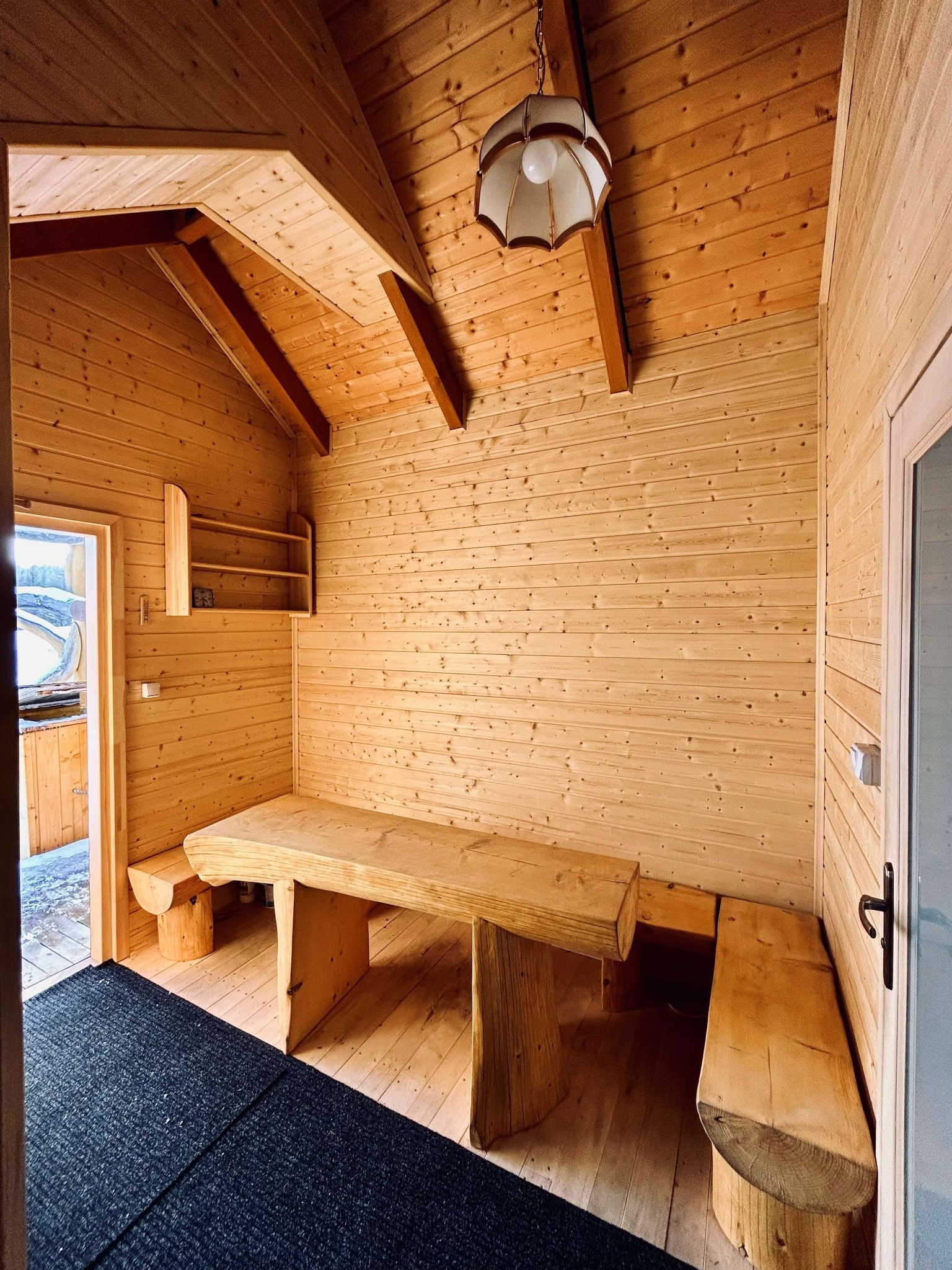 Wnętrze drewnianego domku z surowym stołem i ławkami z bali, widok na zewnątrz przez otwarte drzwi.