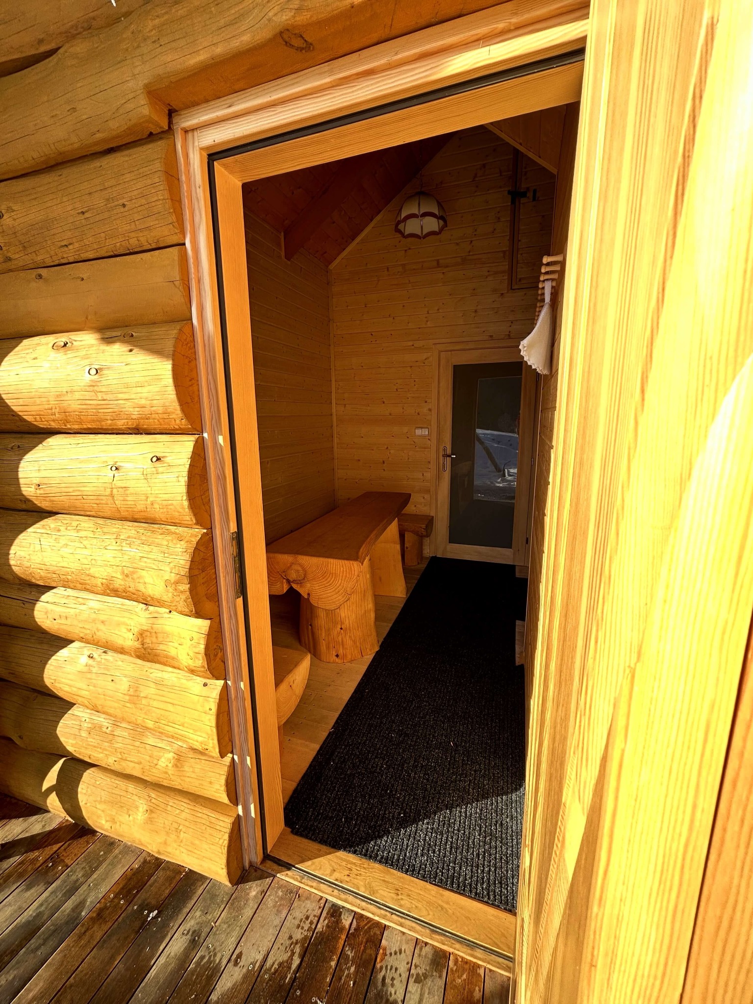 Wnętrze drewnianego domku z bali, widok z zewnątrz przez otwarte drzwi, z widoczną ławką z pnia drzewa i czarnym dywanikiem przy wejściu.