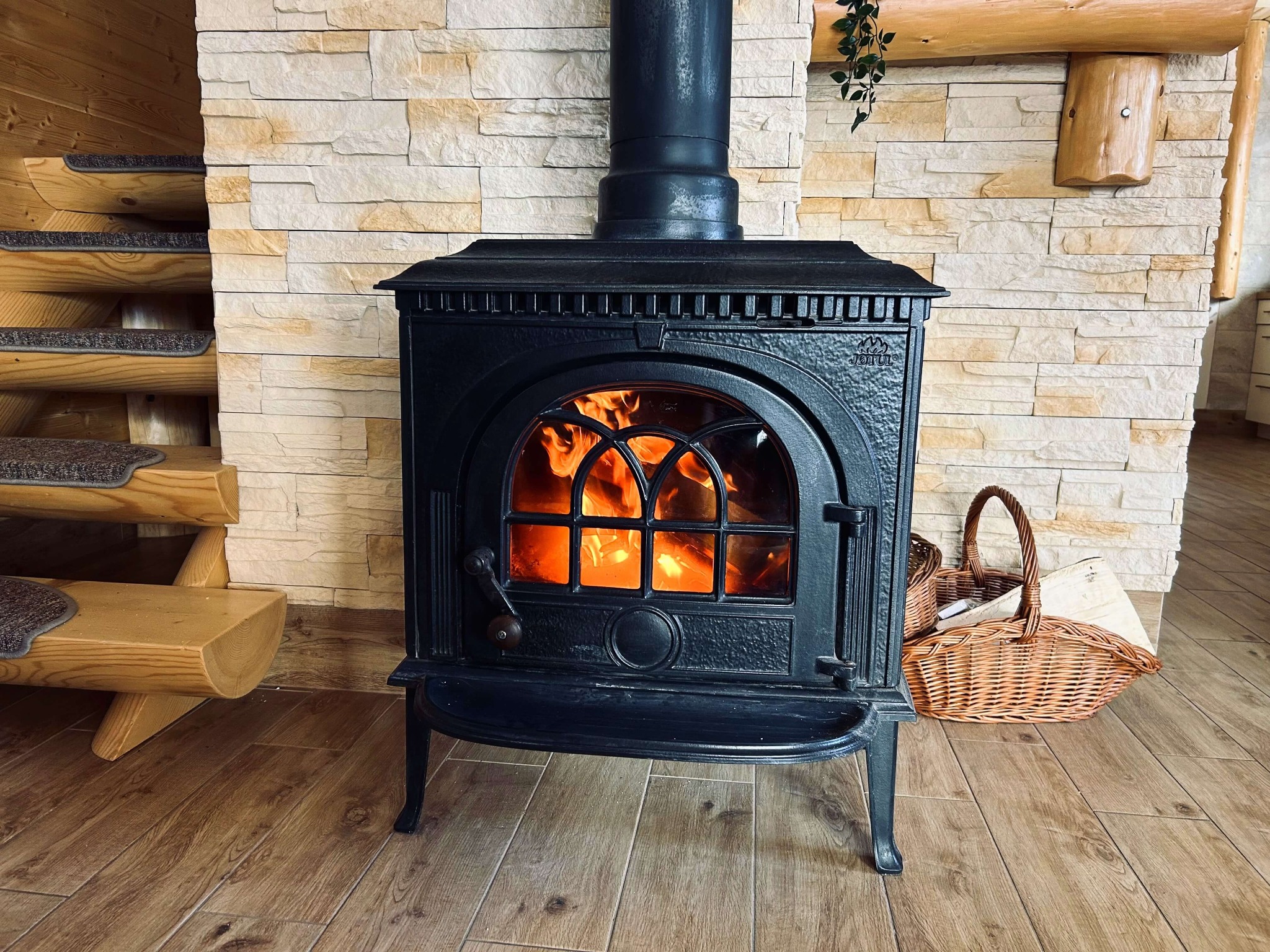 Czarny piec kominkowy z płomieniami za szybą, ustawiony na drewnianej podłodze w rustykalnym wnętrzu z kamienną ścianą i drewnianymi schodami.