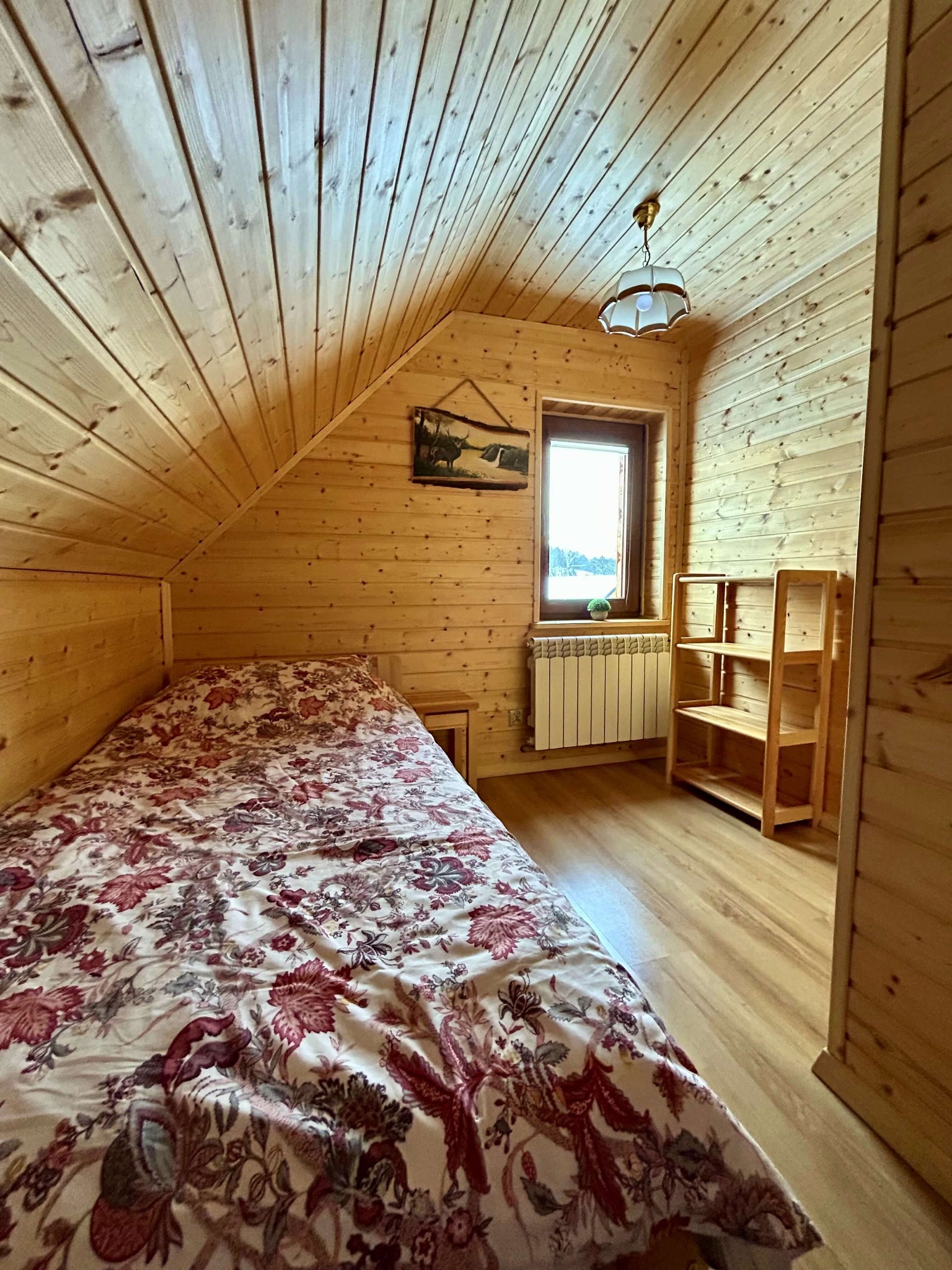 Pokój w stylu rustykalnym z drewnianymi ścianami i skośnym sufitem, łóżko z kwiecistą pościelą, drewniany regał i okno z widokiem na zewnątrz.