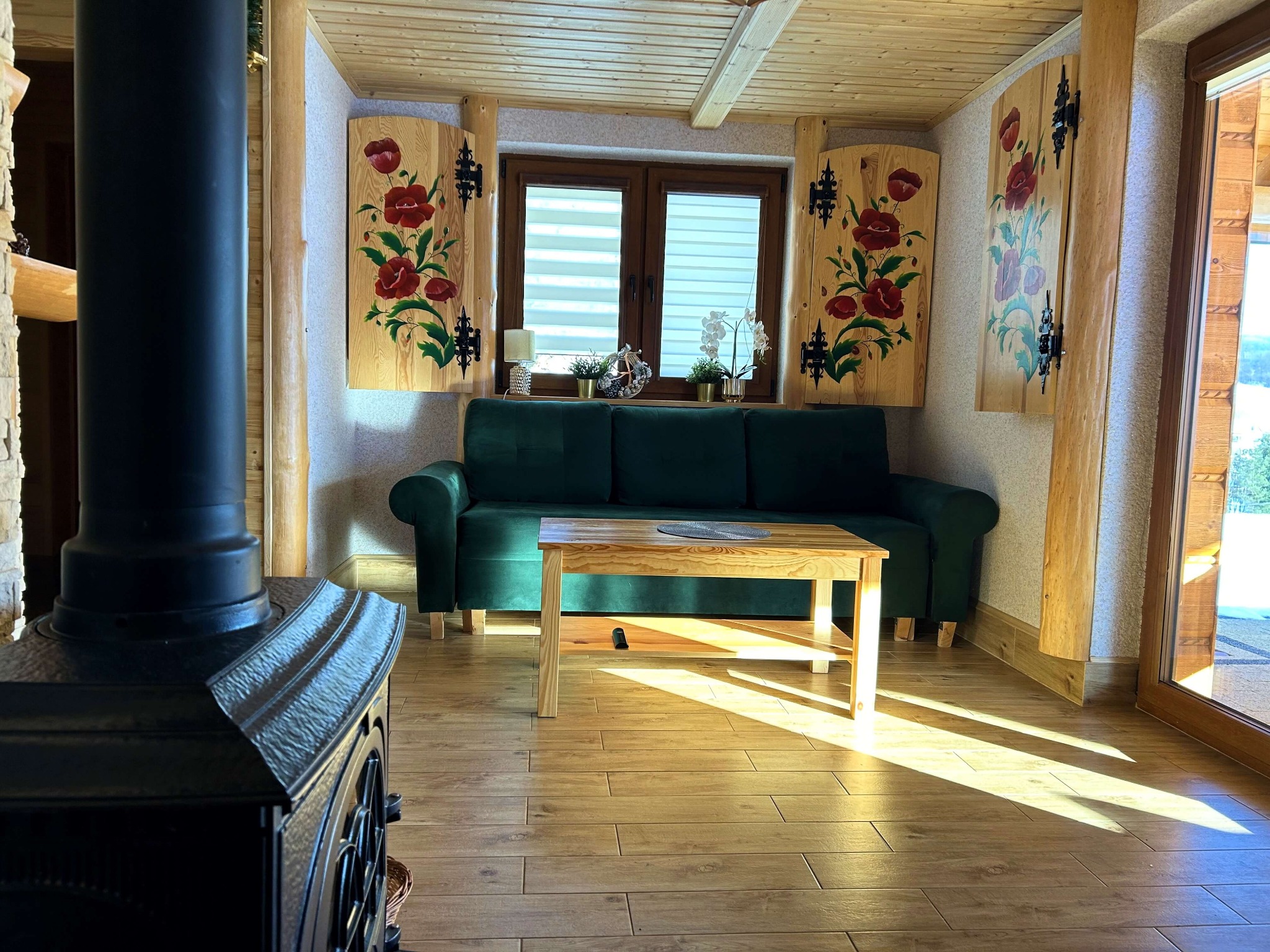 Rustykalny salon z zieloną sofą, drewnianym stolikiem kawowym, piecem kaflowym i okiennicami z motywem maków.