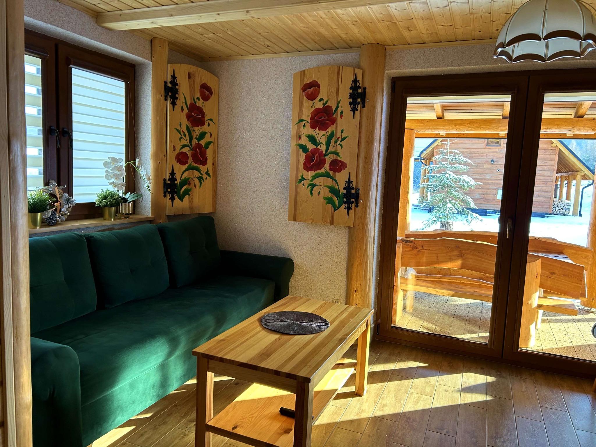 Wnętrze drewnianego domku z zieloną sofą, drewnianym stolikiem kawowym i widokiem na zasypany śniegiem krajobraz za oknem.