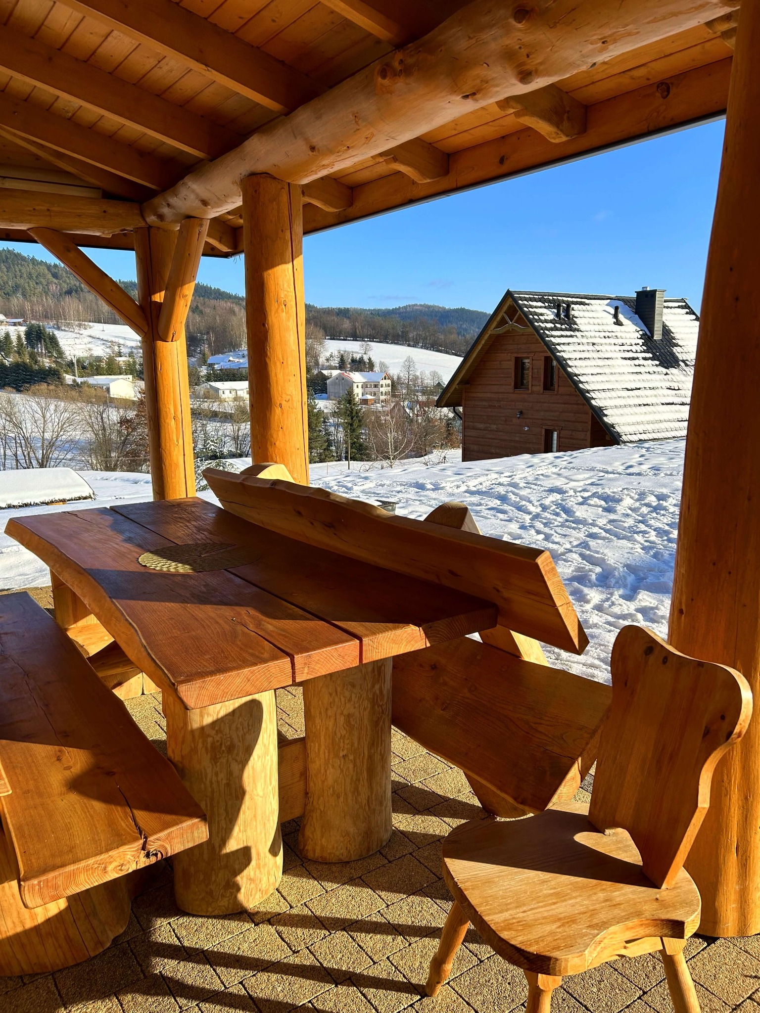 Zadaszony taras z drewnianym stołem i krzesłami z widokiem na ośnieżony krajobraz i drewniany dom w zimowej scenerii.