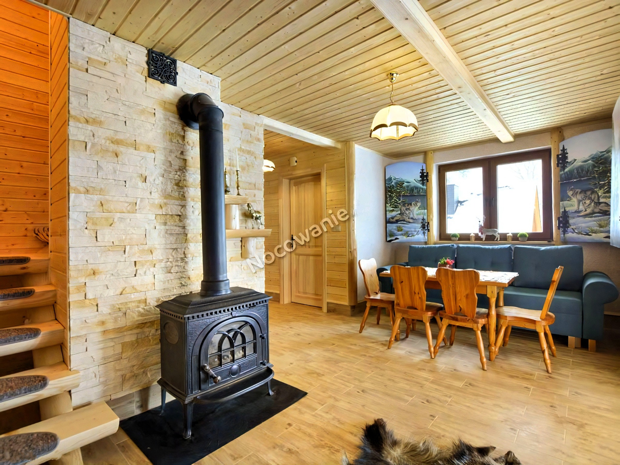 Rustykalne wnętrze: drewniane wykończenie, kamienna ściana z żeliwnym piecem, schody z dywanowymi nakładkami, stół z krzesłami i sofa pod oknem z dekoracyjnymi malowidłami.