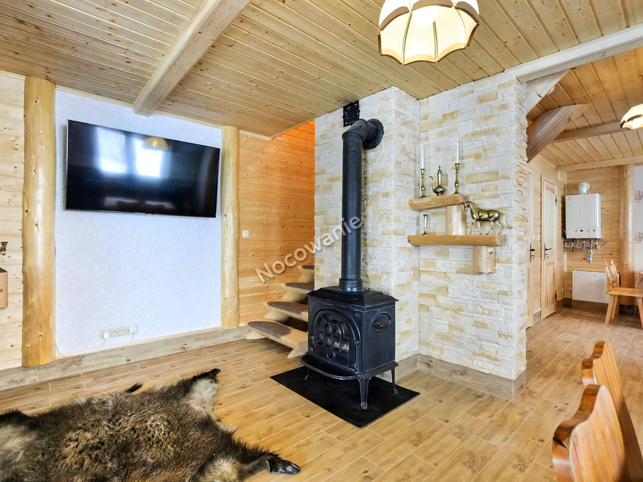 Wnętrze drewnianego domu z kominkiem żeliwnym, skórzaną skórą na podłodze i telewizorem na ścianie, oświetlone lampą wiszącą.