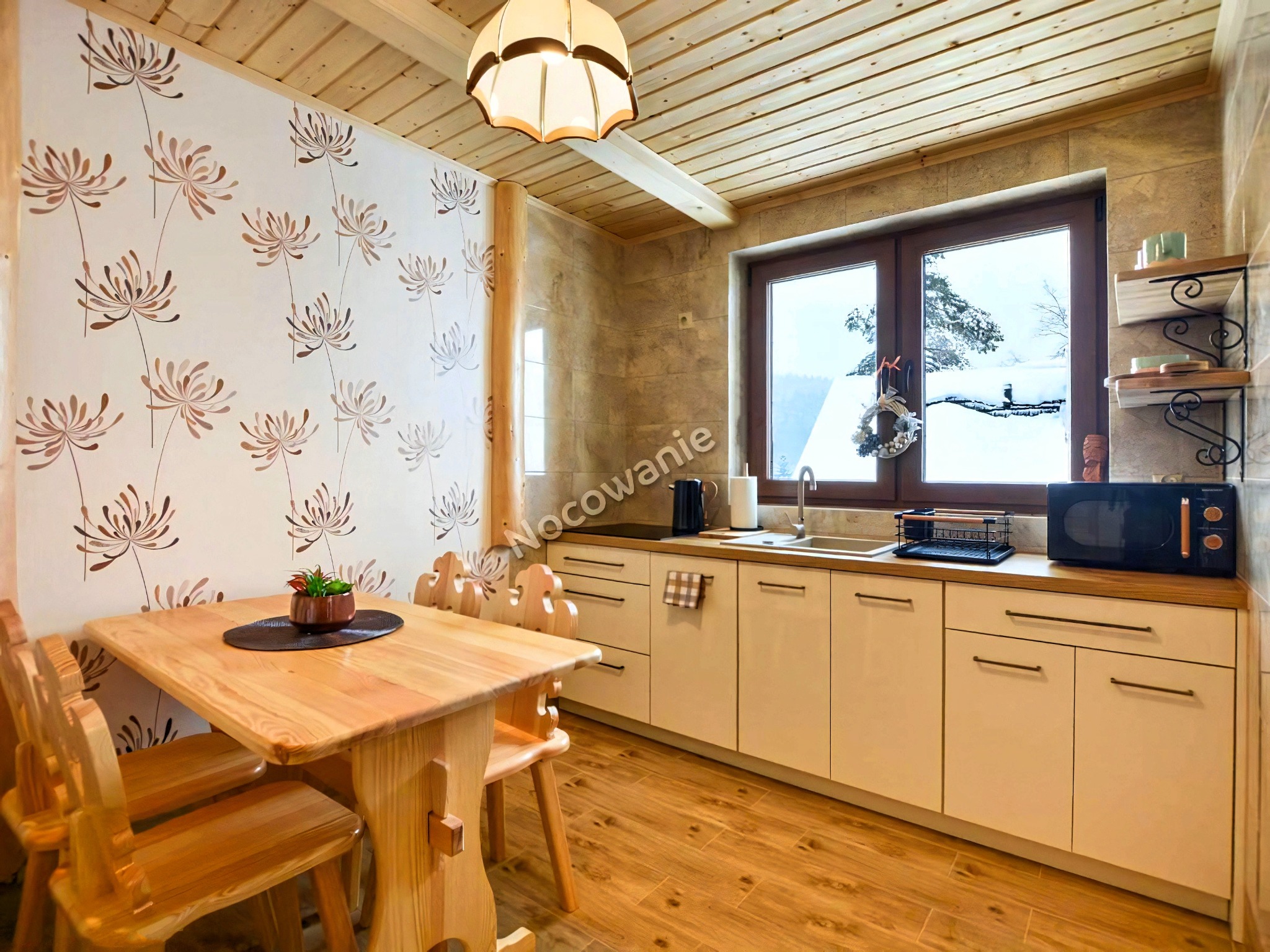 Drewniana kuchnia z widokiem na zaśnieżony krajobraz, stół z kwiatami w doniczce, wzorzysta tapeta z motywem roślinnym.