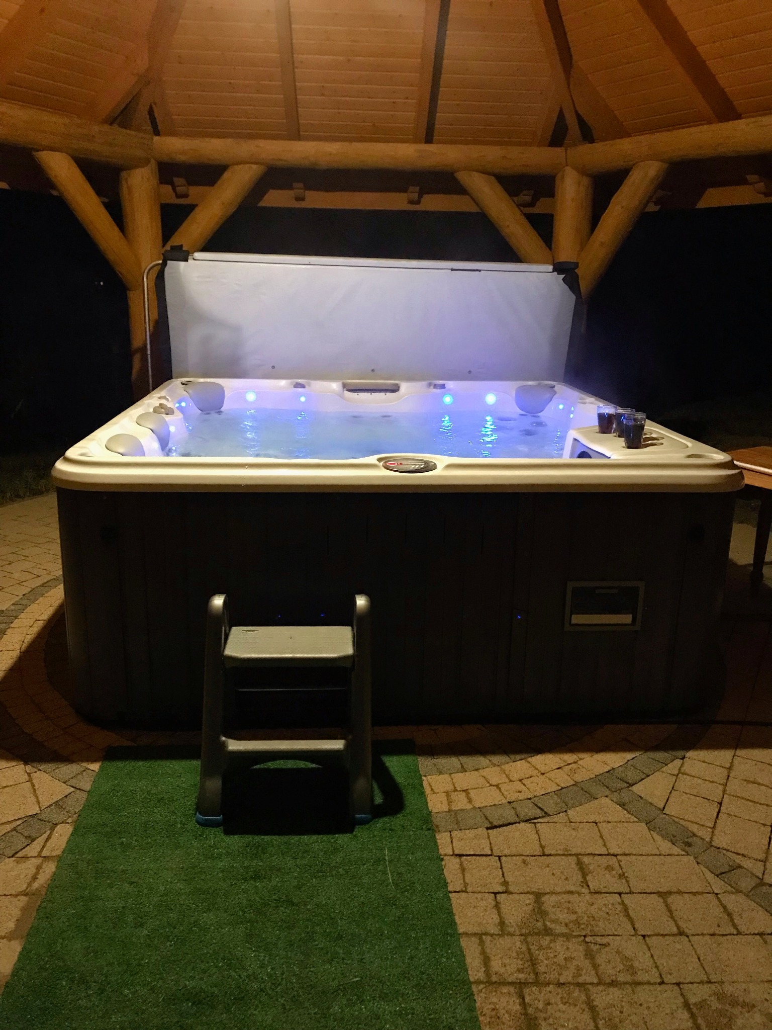 Oświetlone jacuzzi pod drewnianą altaną, z drabinką i sztuczną trawą, wieczorne ujęcie.