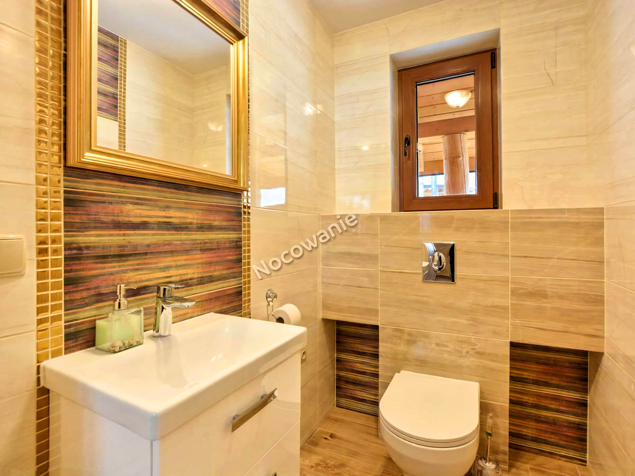 Jasna łazienka z beżowymi płytkami imitującymi drewno, z akcentami w postaci mozaiki i poziomych pasów w odcieniach brązu i złota. Widoczne białe umywalka z szafką, toaleta i lustro w złotej ramie.