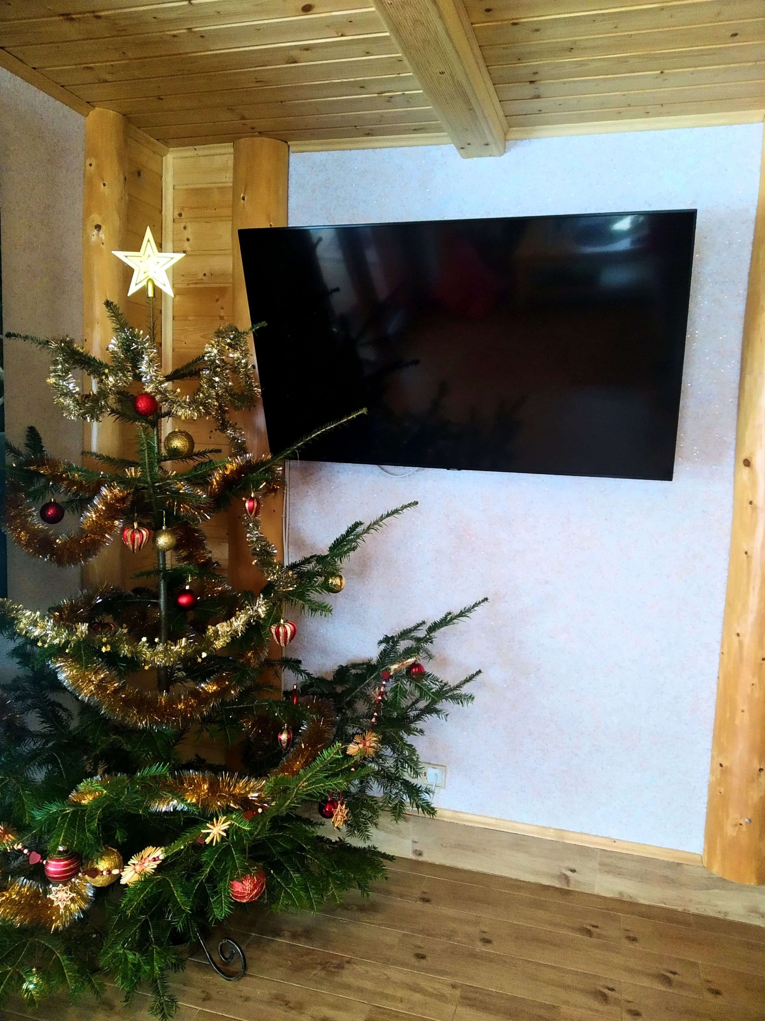Udekorowane drzewko świąteczne z gwiazdą na czubku, obok wiszącego telewizora w drewnianym domku.