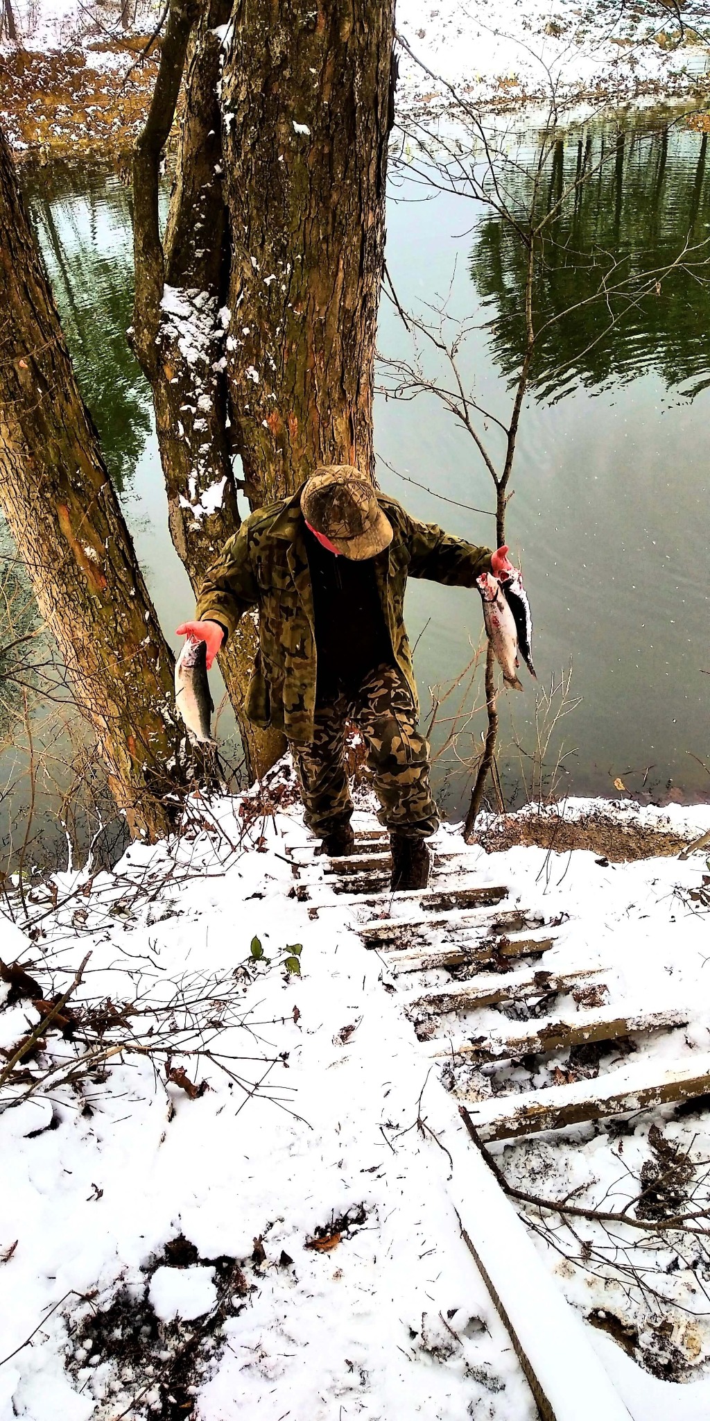 Mężczyzna w stroju kamuflażowym schodzi po schodach w śniegu, trzymając w rękach złowione ryby, z widokiem na rzekę i drzewa.