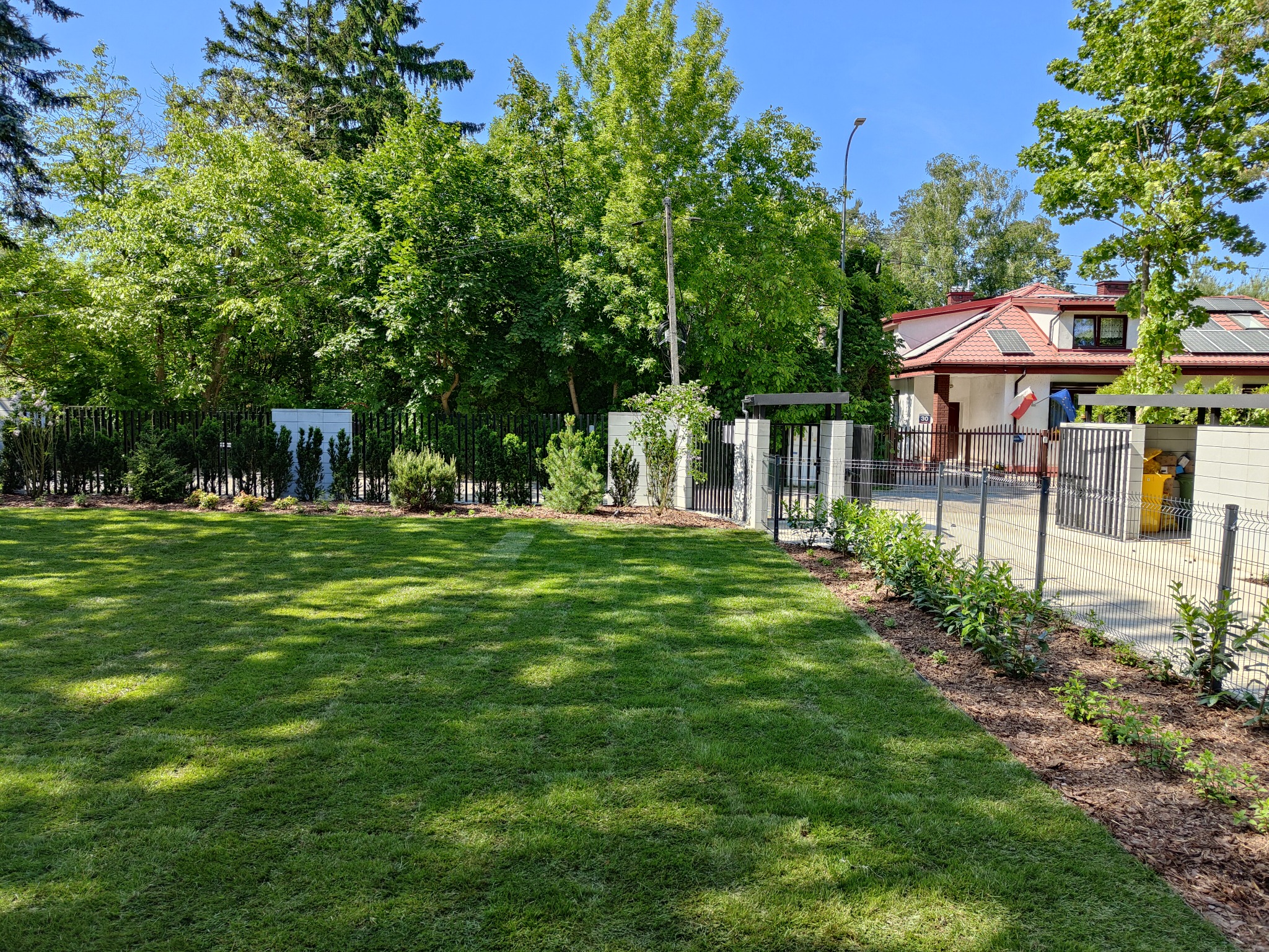 Świeżo założony trawnik z rolki, widoczny pas po wałowaniu, obrzeża wykończone korą, w tle ogrodzenie posesji i budynek mieszkalny z panelami słonecznymi.