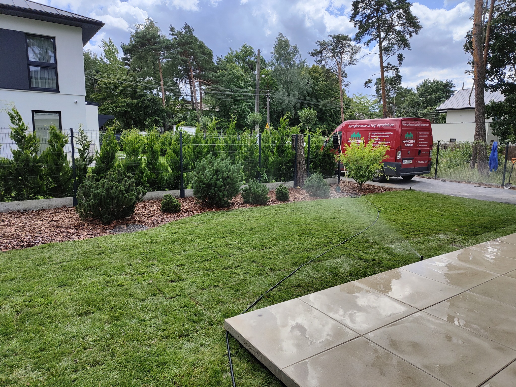 Świeżo założony trawnik z rolki, nawadniany przez automatyczny system zraszający w ogrodzie przy nowoczesnym domu, widoczny samochód firmy ogrodniczej z logo i danymi kontaktowymi.
