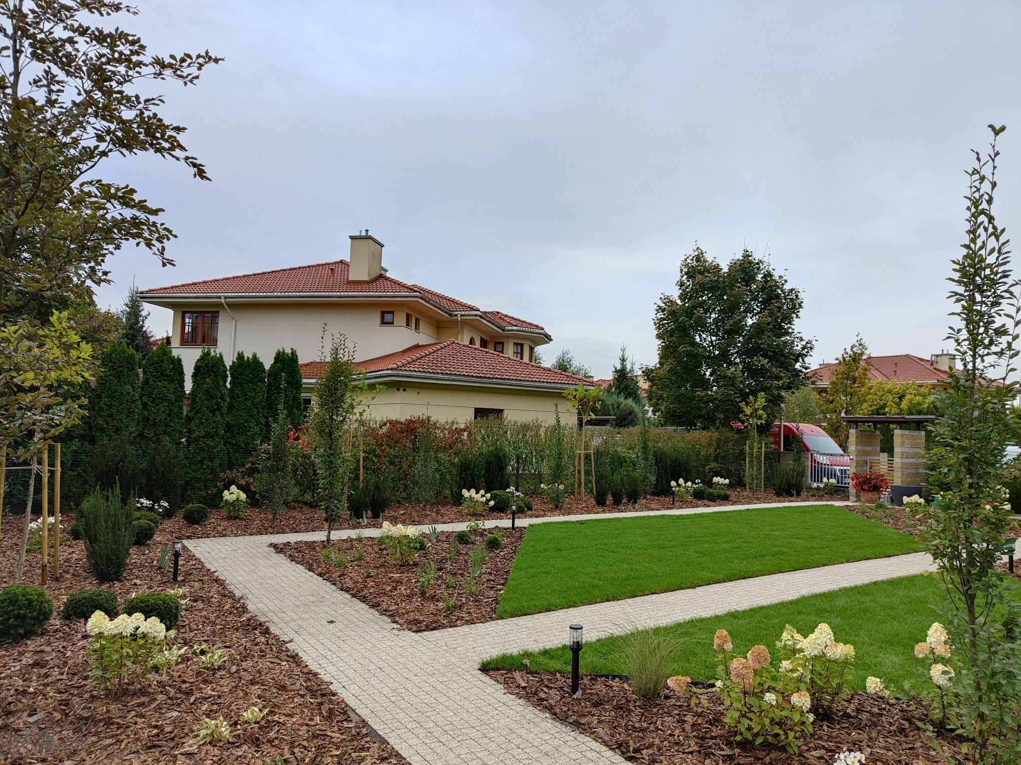 Starannie zaprojektowany ogród przydomowy z równo przystrzyżonym trawnikiem, ścieżkami z kostki brukowej, rabatami z korą i kwitnącymi hortensjami na tle domu z czerwoną dachówką i zielenią drzew.