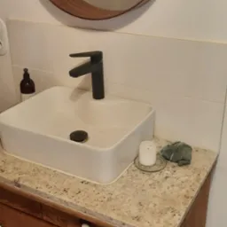 Nowoczesna łazienka z kwadratową umywalką, czarną armaturą i blatem z jasnego granitu, uzupełniona okrągłym lustrem w drewnianej ramie i świecą. Szafka pod umywalką w stylu rustykalnym...