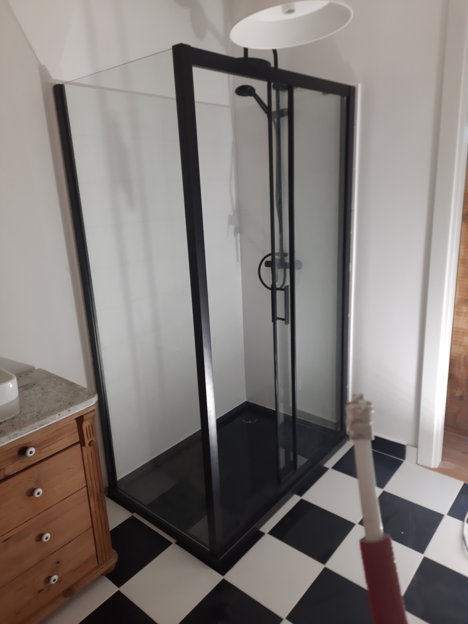 Nowoczesna kabina prysznicowa z czarnymi ramami i armaturą, na tle białej ściany i czarno-białej szachownicy podłogowej, widoczny fragment drewnianej komody z marmurowym blatem i narzędzie...