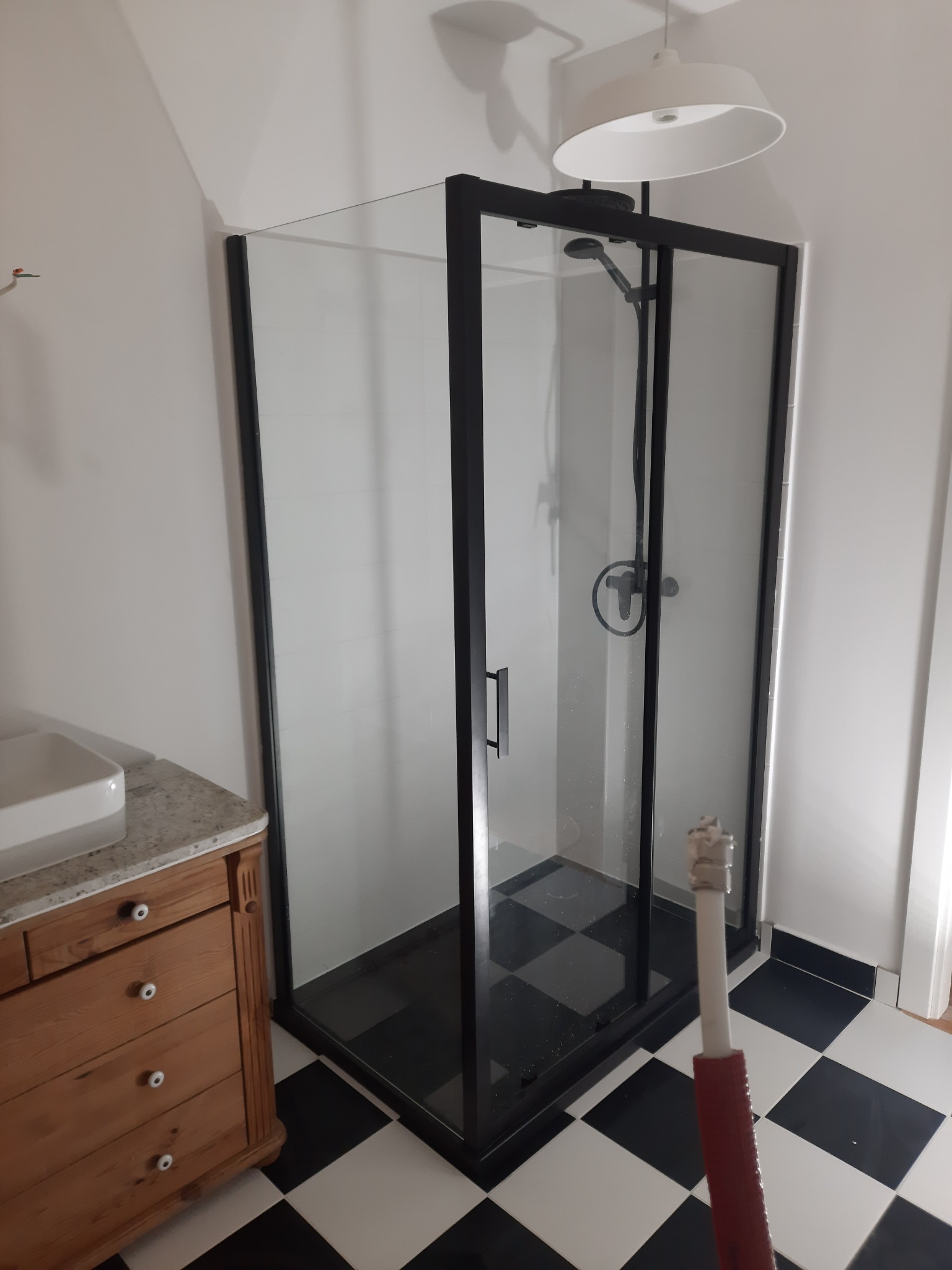 Nowoczesna kabina prysznicowa z czarną ramą i szklanymi ściankami na czarno-białej podłodze w szachownicę, widoczny fragment drewnianej komody z umywalką i biała lampa sufitowa.