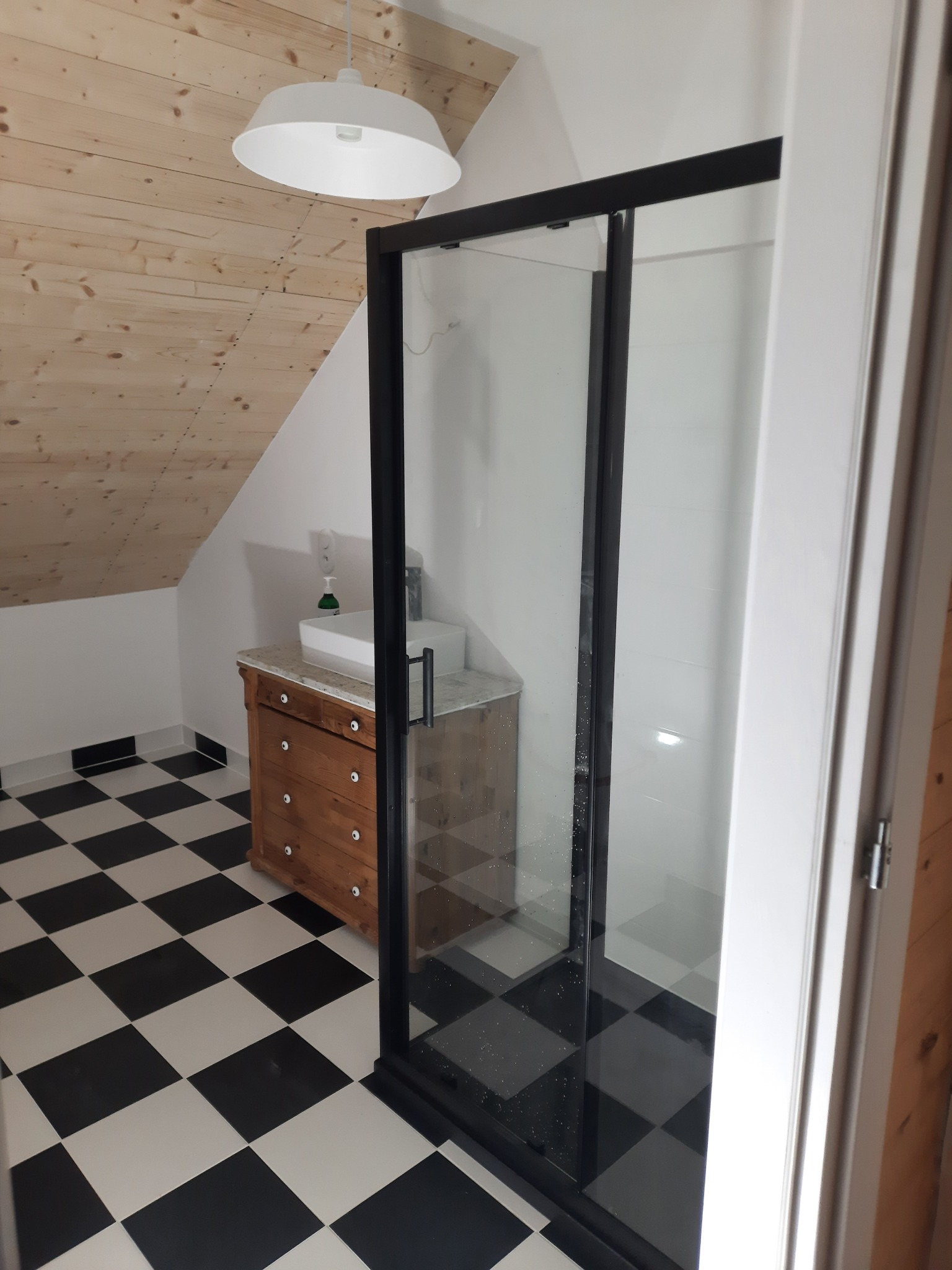 Nowoczesna łazienka na poddaszu z kabiną prysznicową z czarną ramą, umywalką na drewnianej komodzie i białą lampą wiszącą, wykończona w czarno-białą szachownicę.