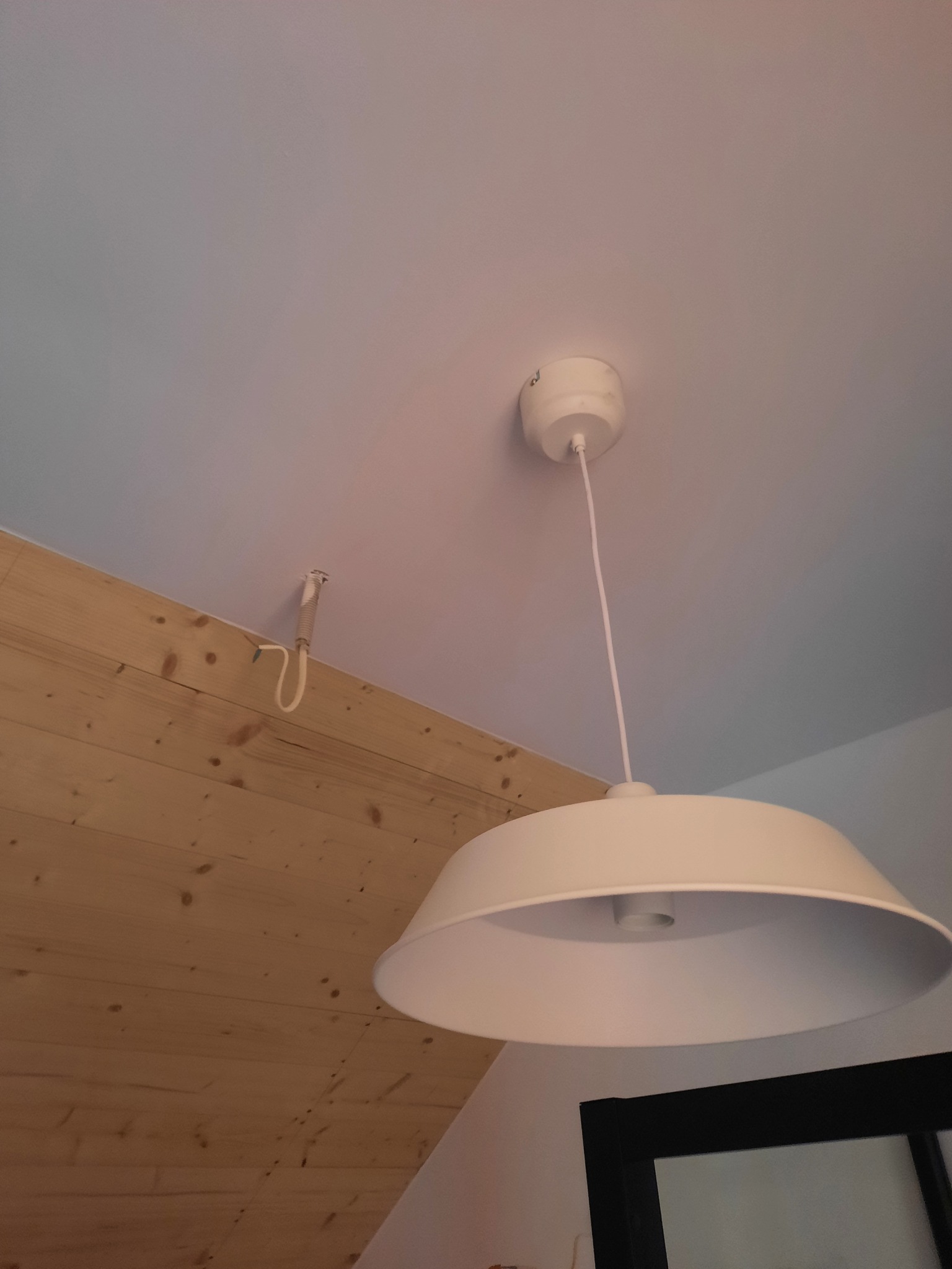 Widok z dołu na białą lampę wiszącą, zamontowaną na białym suficie, z widoczną drewnianą boazerią na ścianie i fragmentem czarnej ramy lustra w tle.