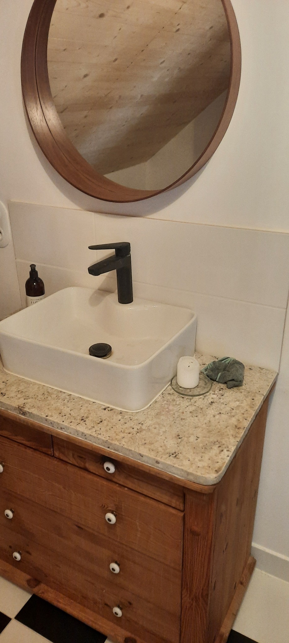 Nowoczesna łazienka z kwadratową umywalką, czarną armaturą i blatem z jasnego granitu, uzupełniona okrągłym lustrem w drewnianej ramie i świecą. Szafka pod umywalką w stylu rustykalnym...