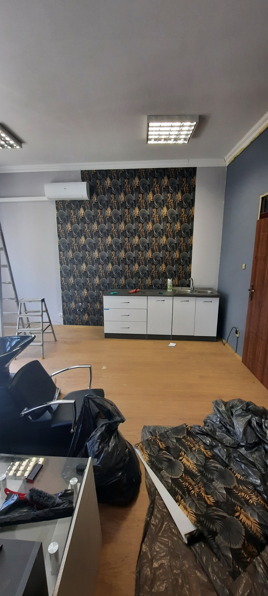 Wnętrze biura w trakcie tapetowania ściany wzorzystą tapetą, widoczne narzędzia, drabina, fragmenty mebli i resztki tapety na podłodze.