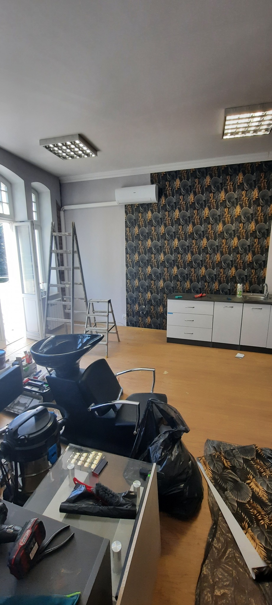 Wnętrze salonu fryzjerskiego w trakcie prac: położona tapeta w liście, drabiny, umywalka fryzjerska, meble i narzędzia na podłodze.