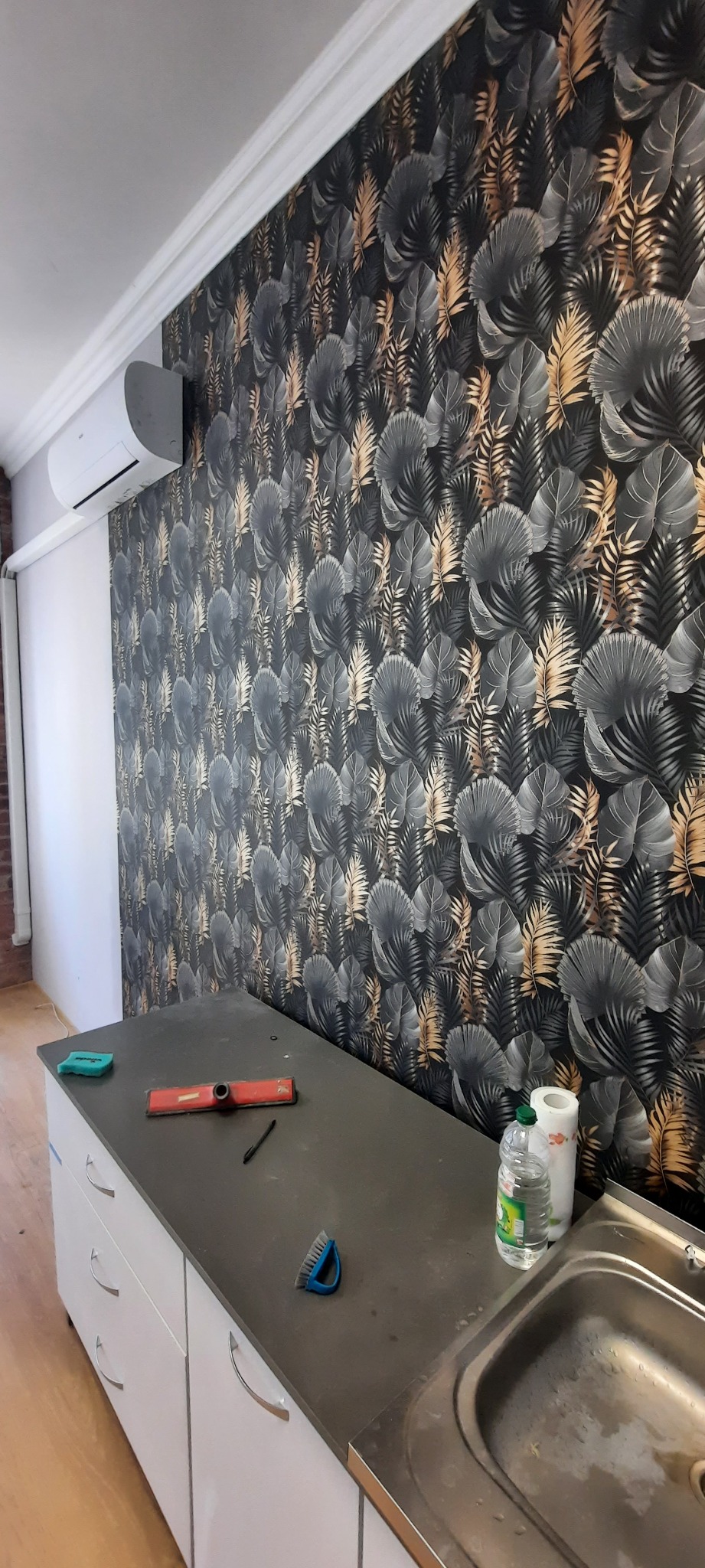 Ściana pokryta tapetą w motywy liściaste w odcieniach szarości i złota, z widocznym poziomem, ołówkiem i szczotką na blacie, sugerując prace wykończeniowe.