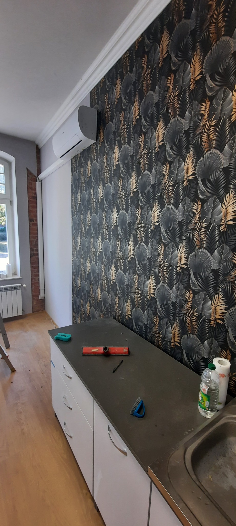 Pokój po remoncie: ściana z tapetą w motywy roślinne, biały klimatyzator, białe szafki kuchenne, poziomica i pędzel na blacie.