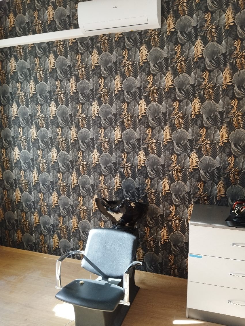 Ściana w salonie fryzjerskim pokryta tapetą w motywy liści, z zamontowanym klimatyzatorem Haier u góry i stanowiskiem do mycia włosów z czarną umywalką oraz szarym fotelem na pierwszym planie.