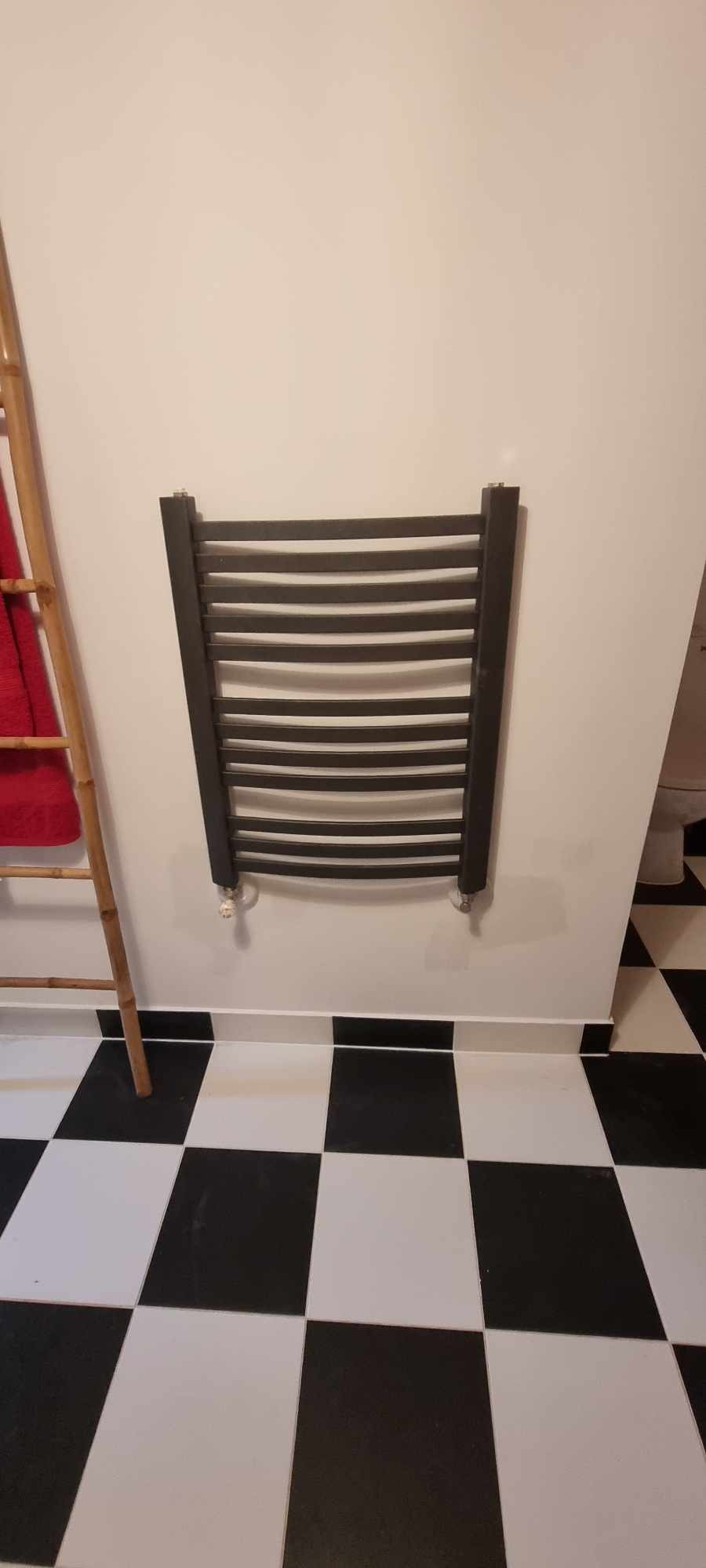 Ciemny, drabinkowy grzejnik łazienkowy zamontowany na białej ścianie, z widoczną bambusową drabinką na ręczniki i czarno-białą szachownicą na podłodze.