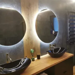 Nowoczesna łazienka z dwoma okrągłymi lustrami podświetlanymi LED, czarnymi umywalkami nablatowymi na drewnianym blacie i szarą okładziną ścienną imitującą kamień.