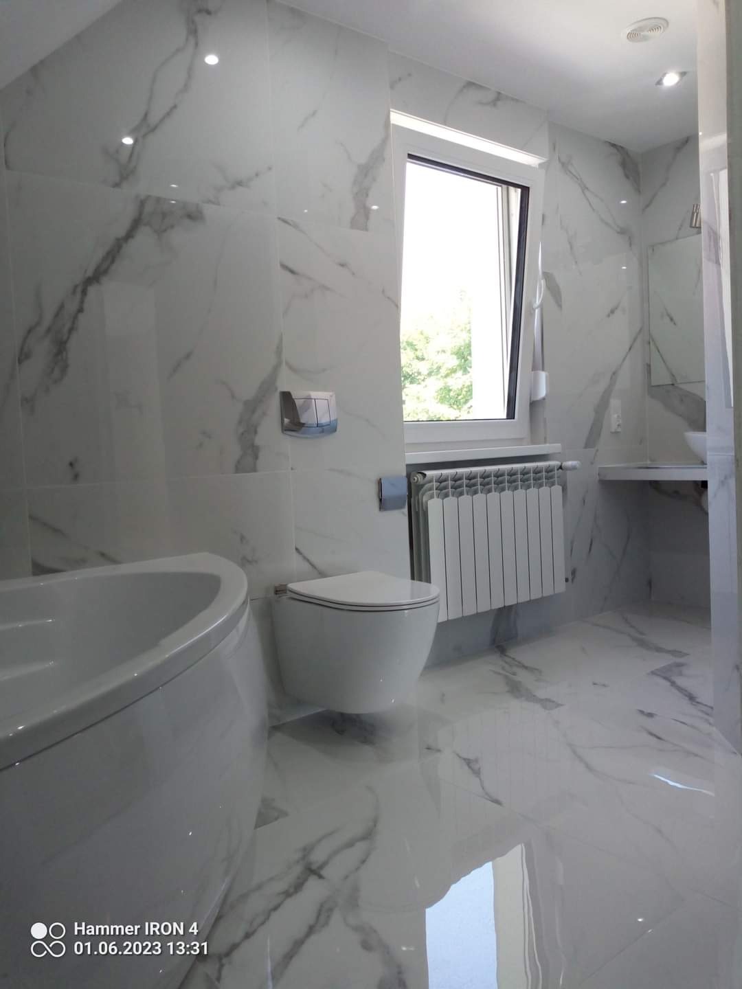 Nowoczesna łazienka wykończona płytkami imitującymi biały marmur z szarymi żyłami, z widoczną wanną, toaletą, grzejnikiem i oknem z widokiem na zieleń.