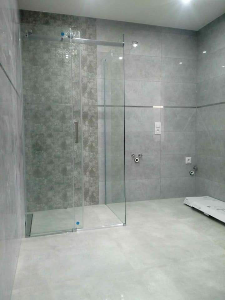 Nowoczesna łazienka z kabiną prysznicową ze szklanymi przesuwnymi drzwiami, szare płytki na ścianach i podłodze, minimalistyczna armatura, widoczne elementy montażowe.