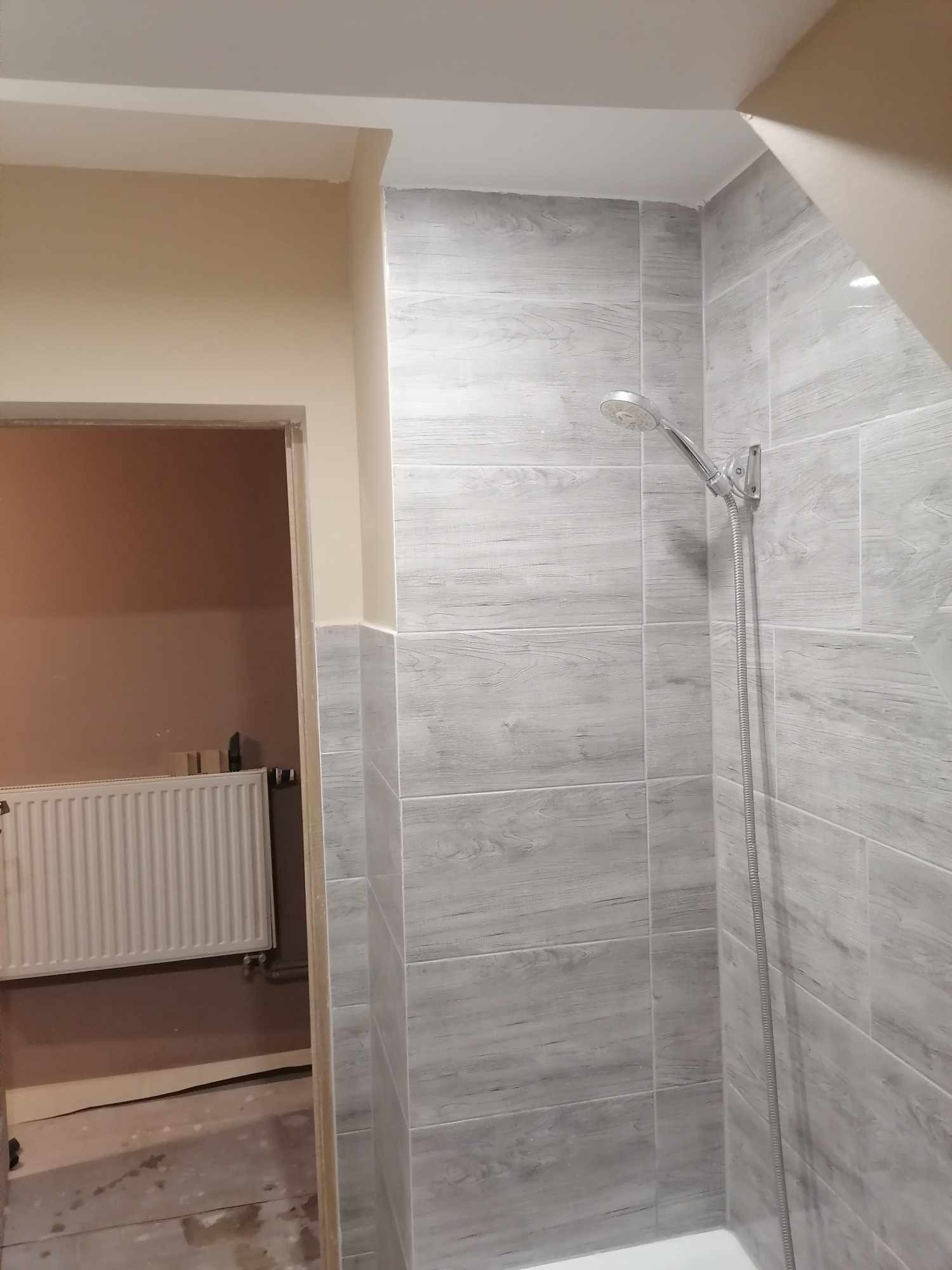 Fragment łazienki w trakcie prac wykończeniowych: szare płytki imitujące drewno na ścianie prysznica, widoczna słuchawka prysznicowa, w tle fragment pokoju z grzejnikiem i niezakończonymi pracami...