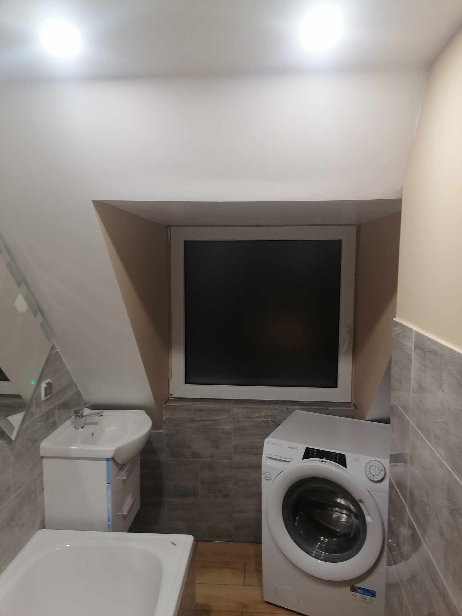 Niewielka łazienka na poddaszu ze skośnym sufitem, wyposażona w białą umywalkę z szafką, wannę i pralkę Candy RapidO. Ściany wyłożone szarą płytką imitującą kamień, okno z czarną szybą.