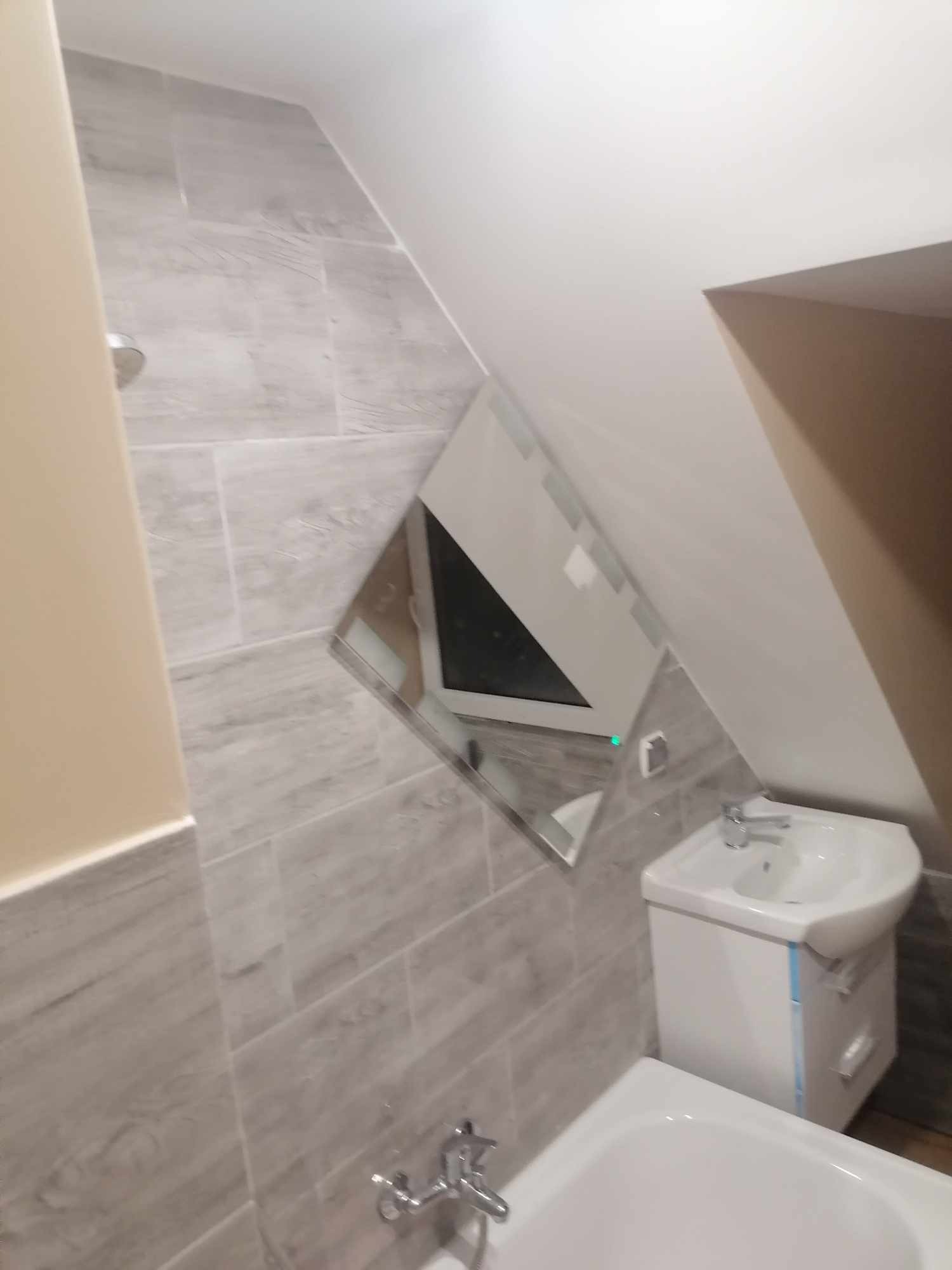 Ukończona łazienka na poddaszu z szarymi płytkami imitującymi drewno, białą umywalką z szafką i wanną, oraz lustrem w kształcie trapezu.