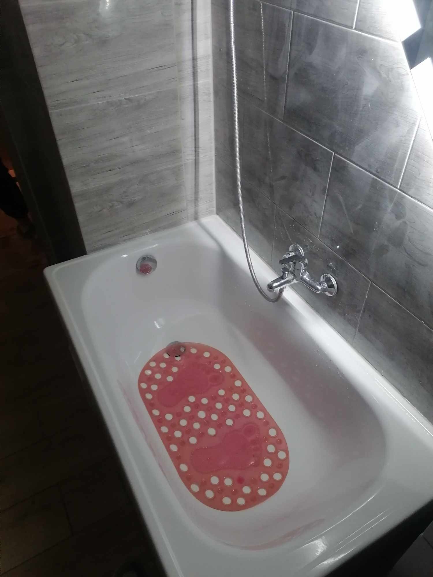 Biała wanna z czerwoną antypoślizgową matą w białe kropki, szare płytki na ścianie i chromowana bateria z wężem prysznicowym.