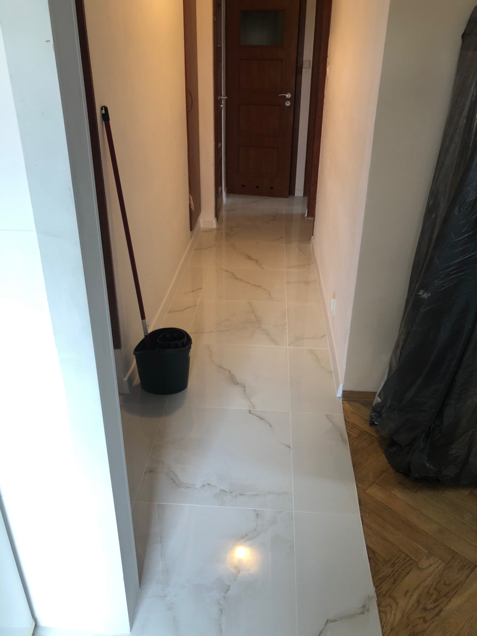 Korytarz po remoncie z nową marmurkową podłogą z płytek, widoczny mop w wiadrze i zabezpieczone folią meble.