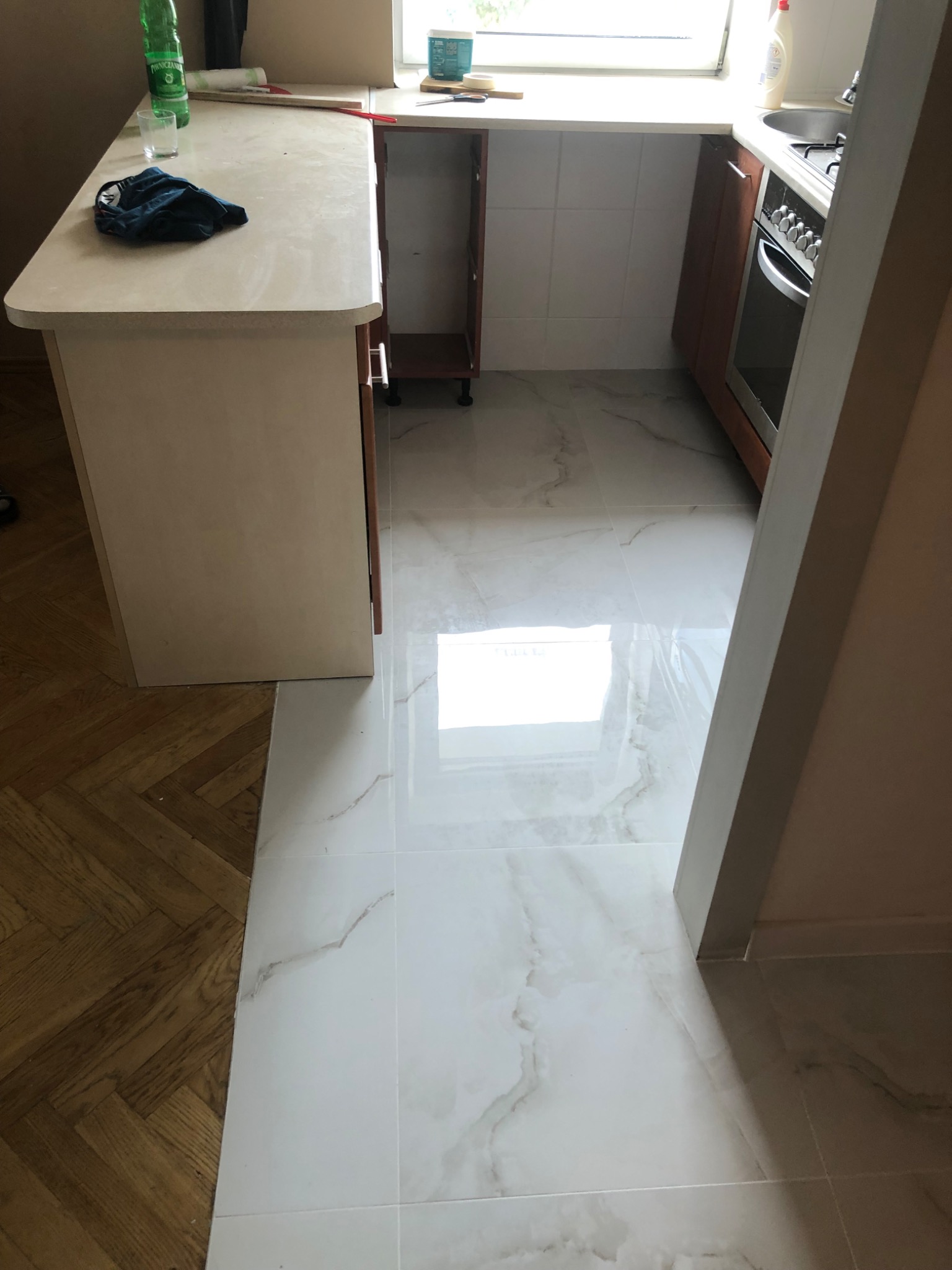 Kuchnia po remoncie z nową białą podłogą z płytek imitujących marmur, kontrastującą z fragmentem starego parkietu i zabudową kuchenną w odcieniach brązu.