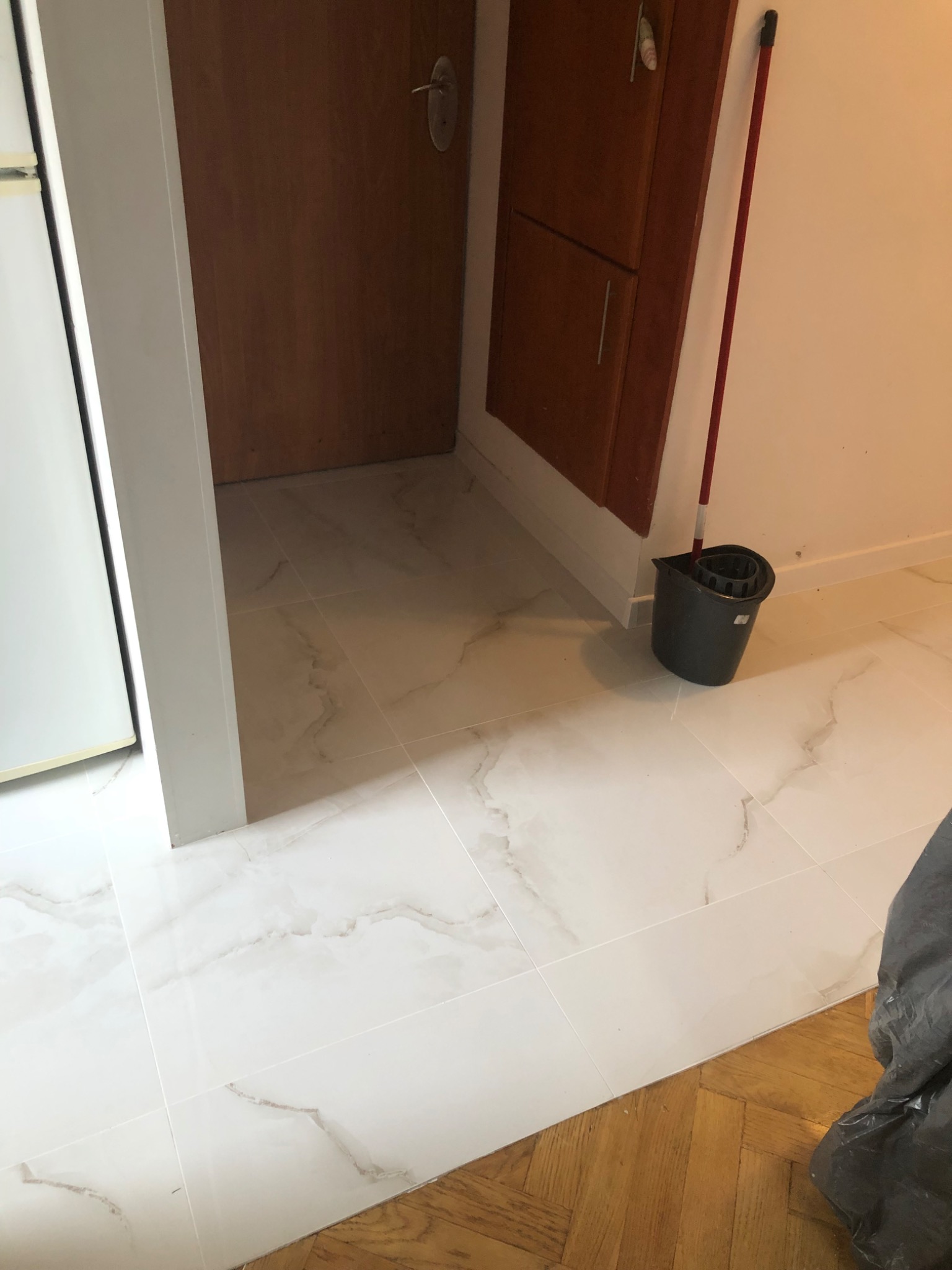 Wykończona podłoga z białych płytek imitujących marmur, widoczny fragment parkietu, wiadro i mop w rogu pomieszczenia.